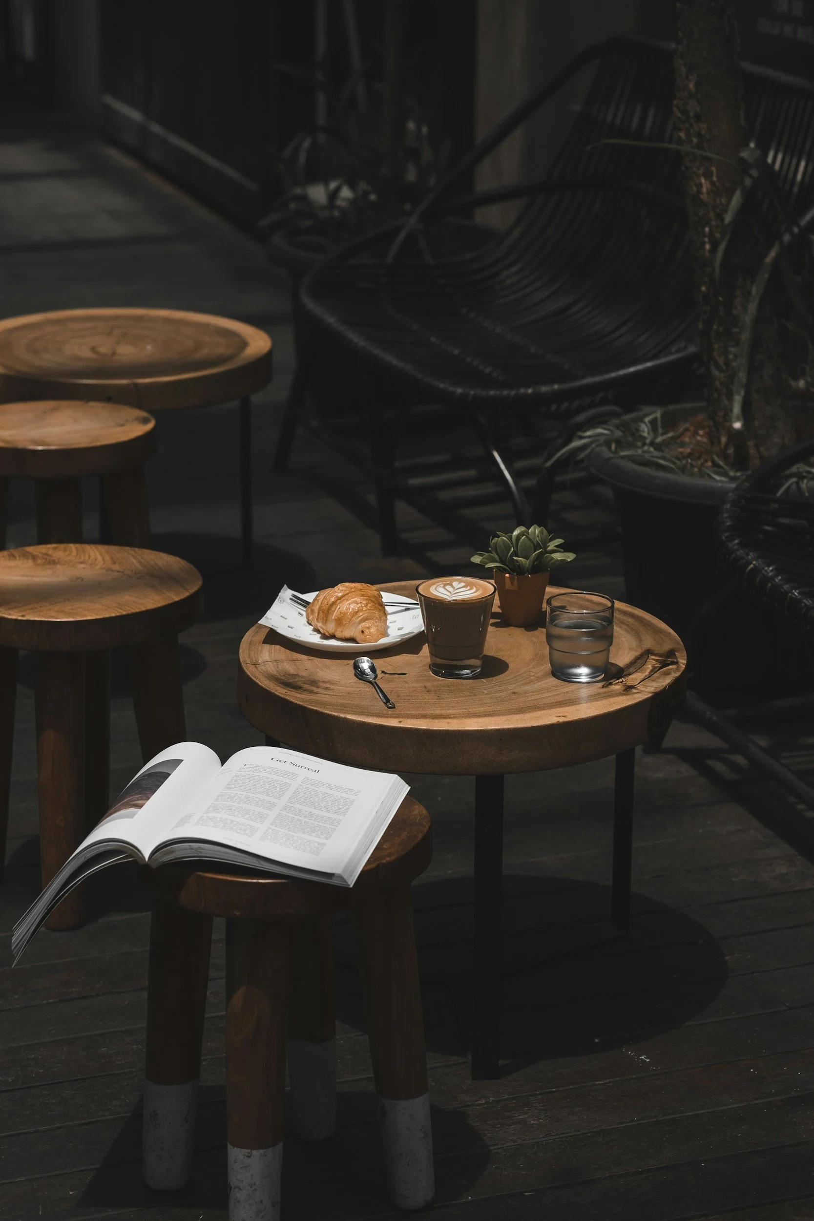 A cozy outdoor café table with a croissant, a latte with latte art, a glass of water, a small potted plant, and an open book on a stool.
