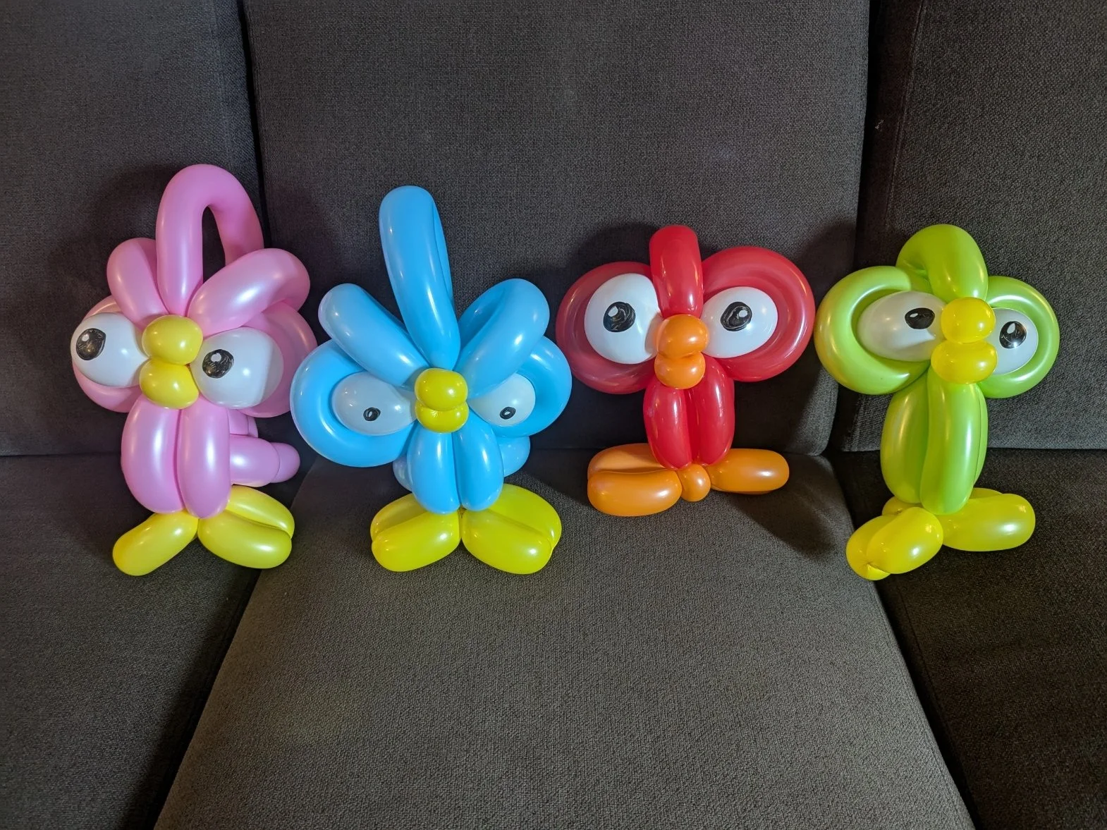 Four balloon animals resembling a pink butterfly, a blue butterfly, a red ladybug, and a yellow butterfly, all with cartoonish eyes, arranged in a row on a dark gray fabric surface.