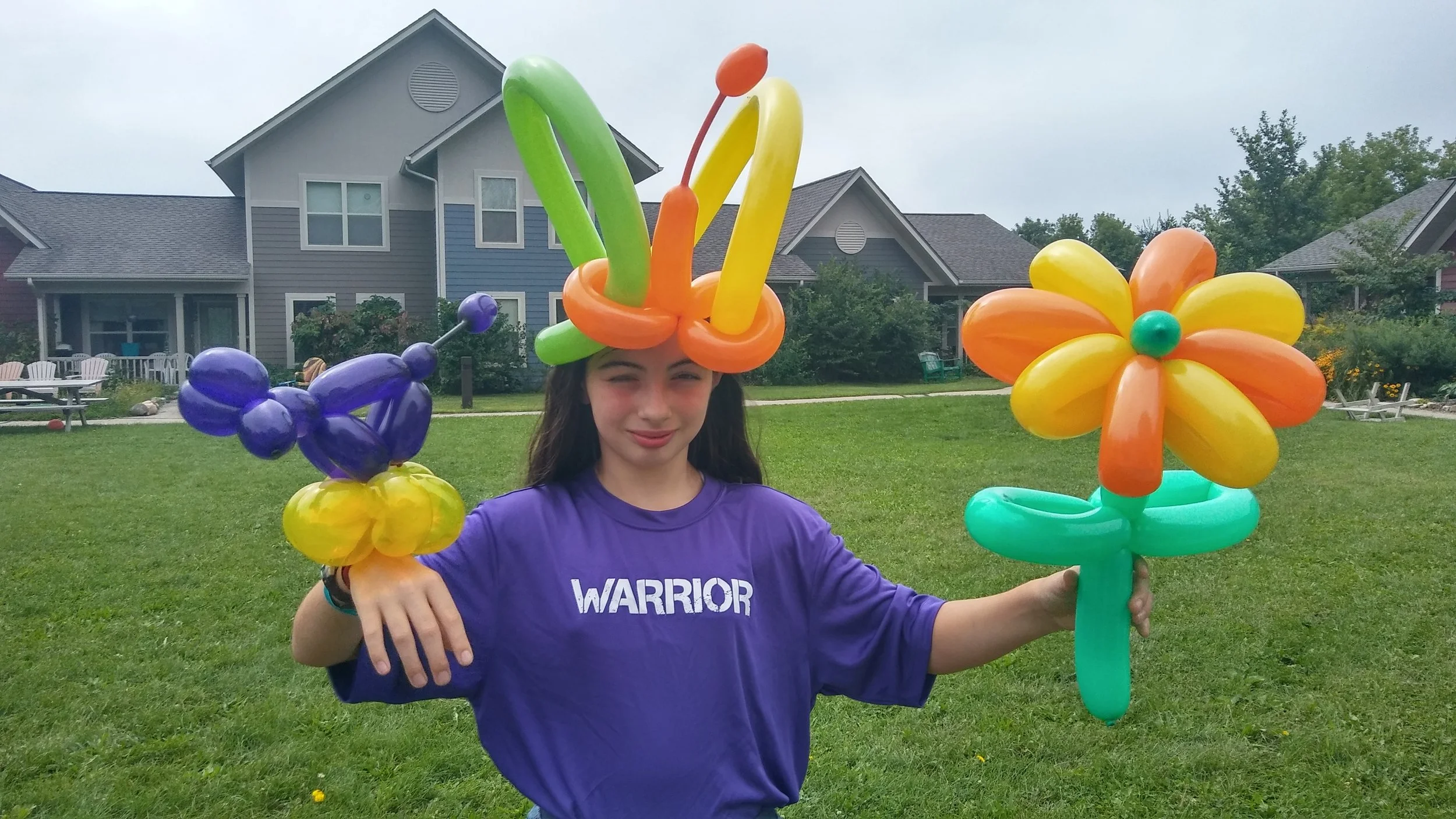 A girl holding colorful balloon sculptures in the shape of a flower, a small helicopter, and a bird, standing on a grassy lawn with houses in the background.