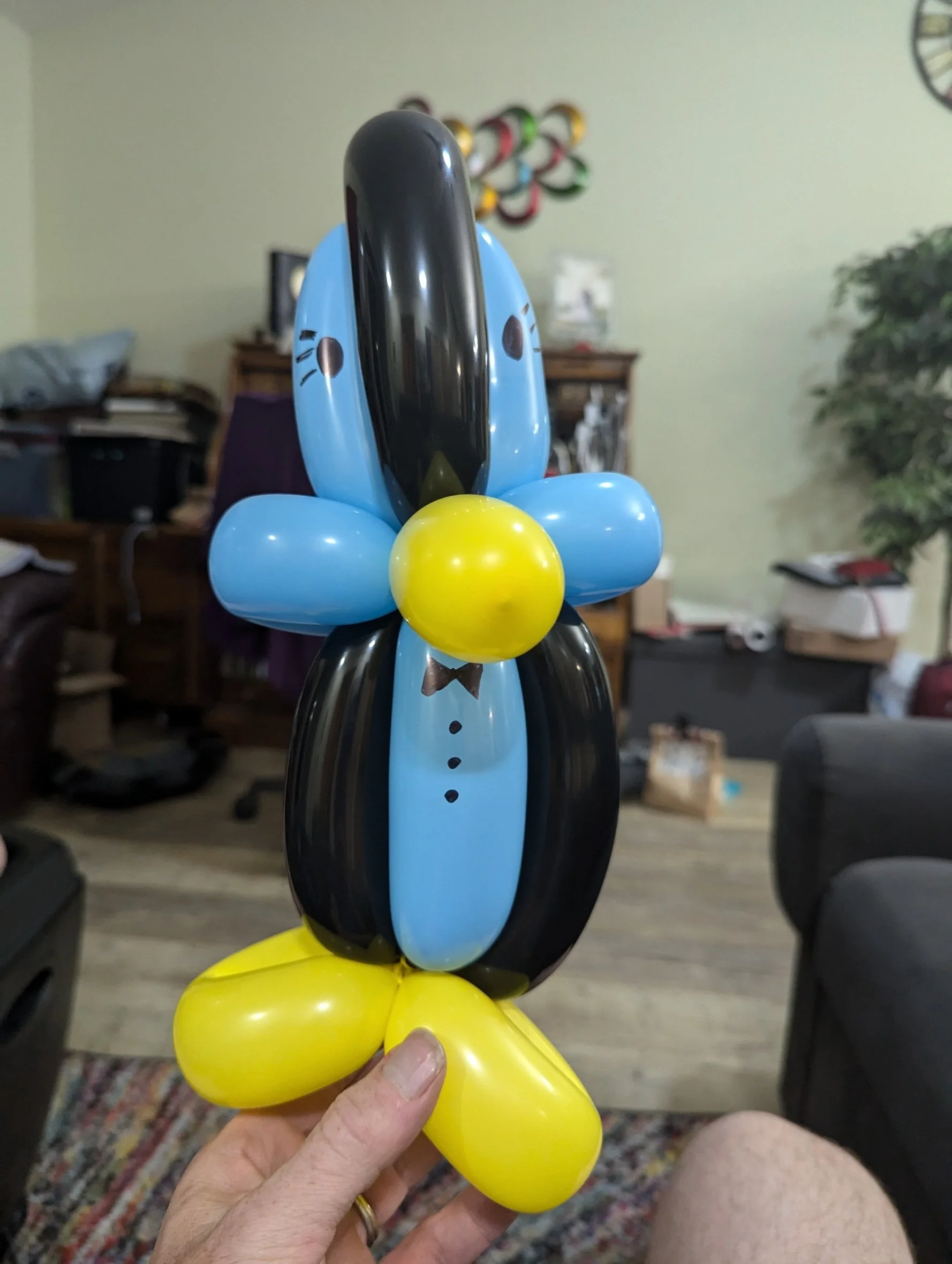 A balloon sculpture of a dog, with yellow, blue, black, and brown balloons, being held by a person with a hand with a gold ring, in a room with a wooden table, chairs, and various objects in the background.