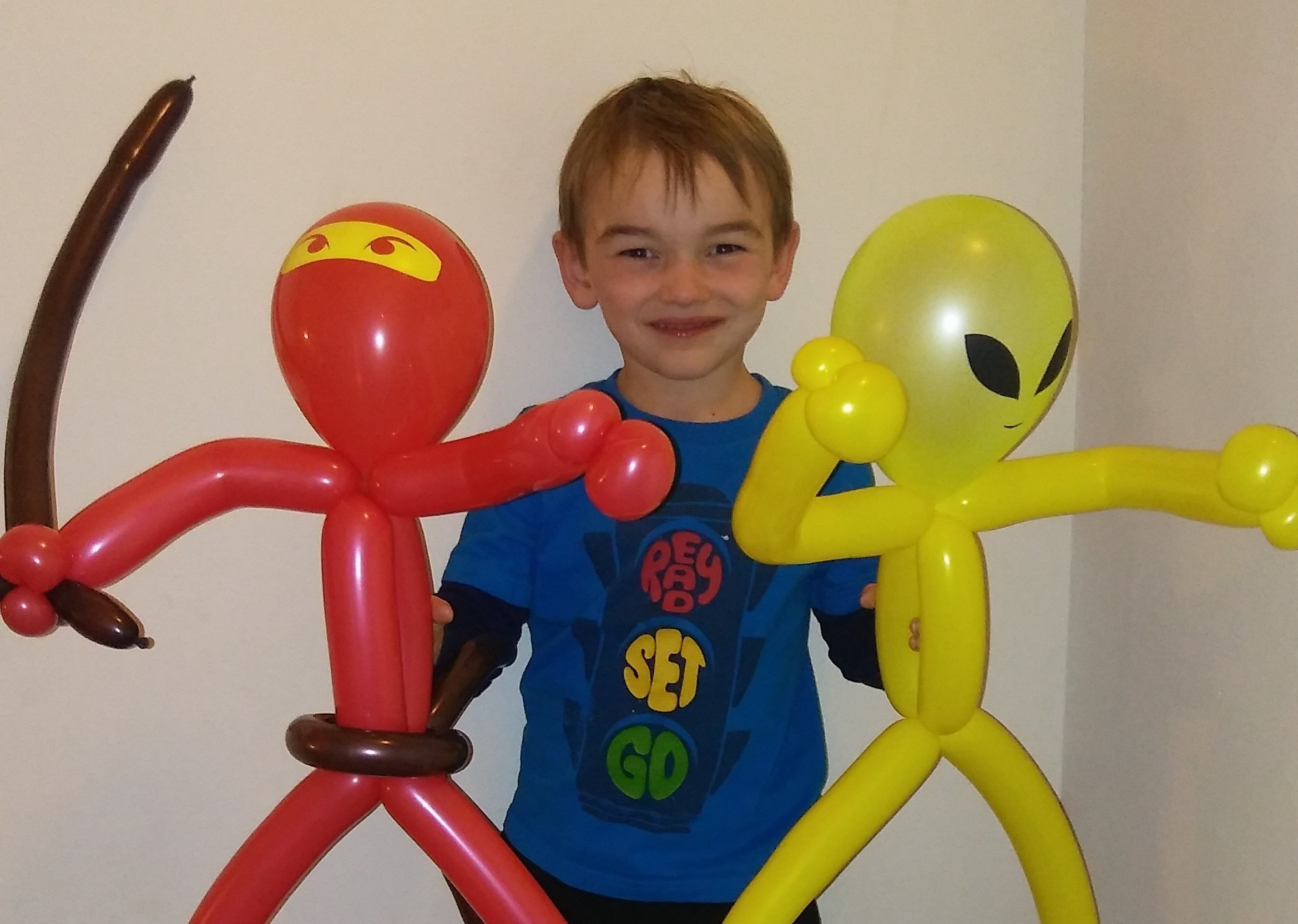 A young boy smiling while holding colorful balloon sculptures of two aliens, one red and one yellow, in front of a plain white wall.