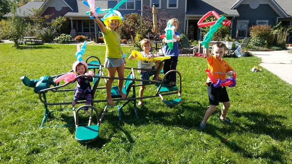 Group of children playing with balloon animals on a playground in a backyard with houses in the background.