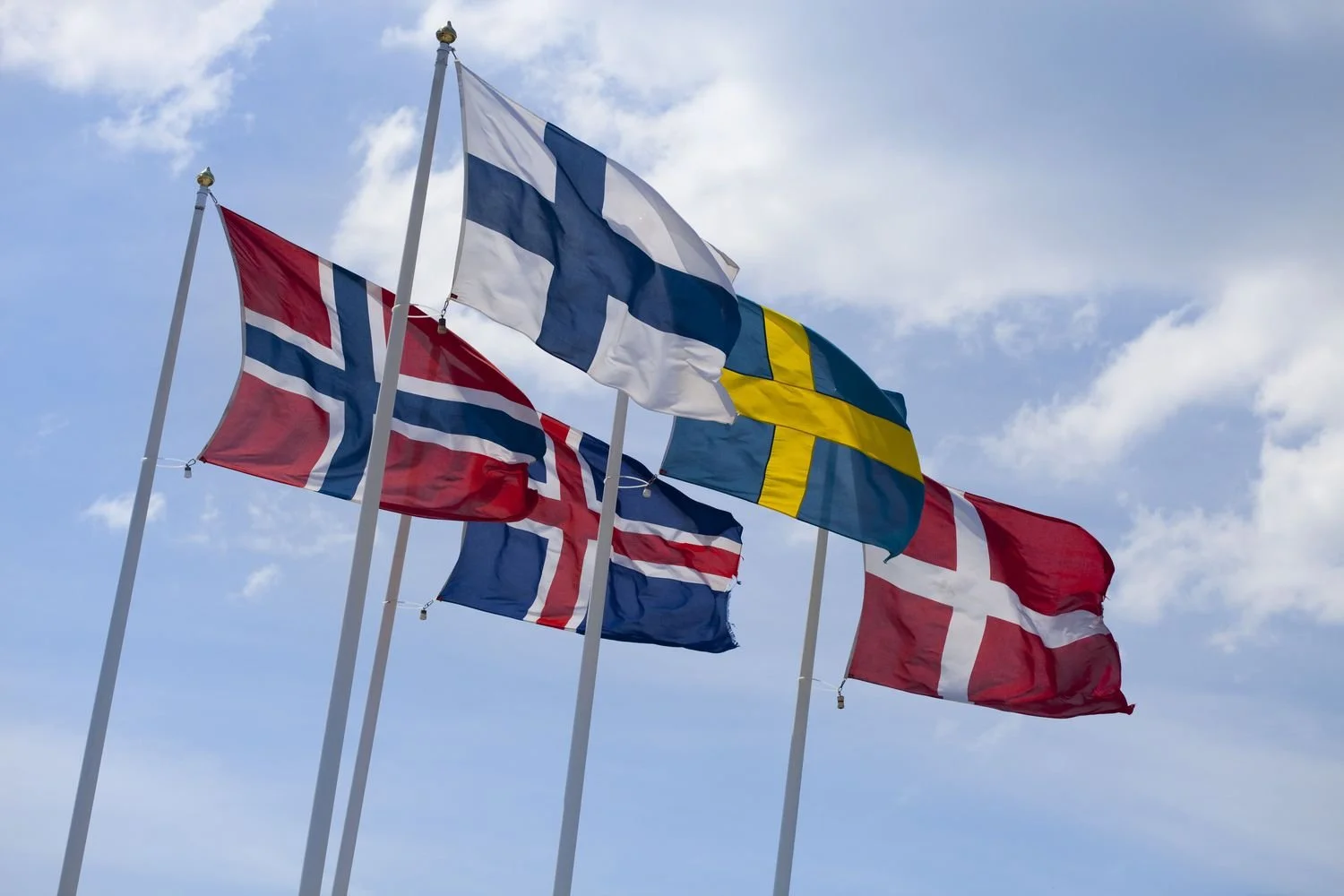 Multiple country flags, including Norway, Finland, Sweden, and Denmark, flying against a partly cloudy sky.