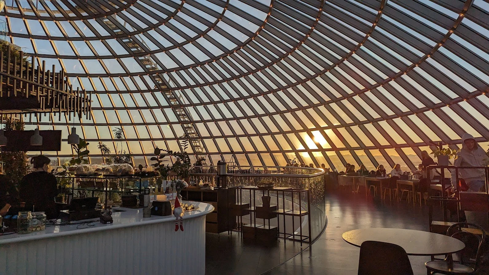 Interior of a modern restaurant or cafe with a curved glass ceiling at sunset, featuring patrons sitting at tables, a marble bar area with staff, and decorative plants.