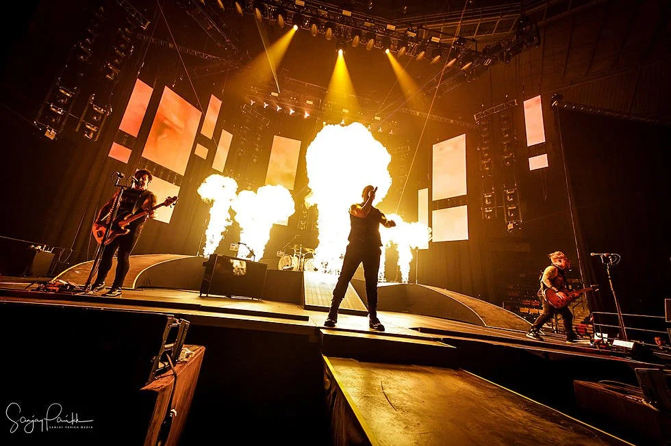 A band performing on stage with flames and large LED screens in the background, illuminated by yellow stage lights.