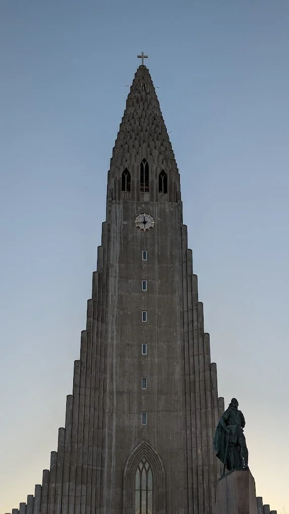Tall stone clock tower with a cross at the top, featuring arched windows near the top, and a statue of a person in front of the base.