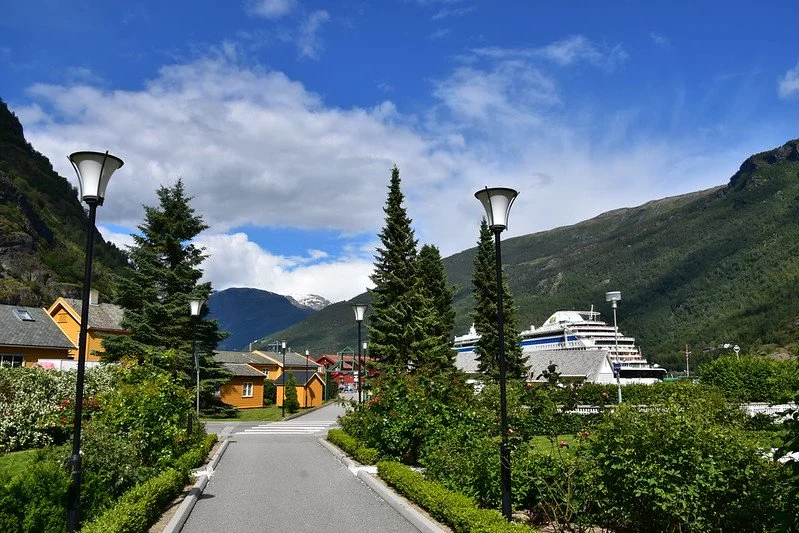 A scenic pathway lined with lamp posts and greenery, with mountains and a cruise ship in the background under a partly cloudy sky.