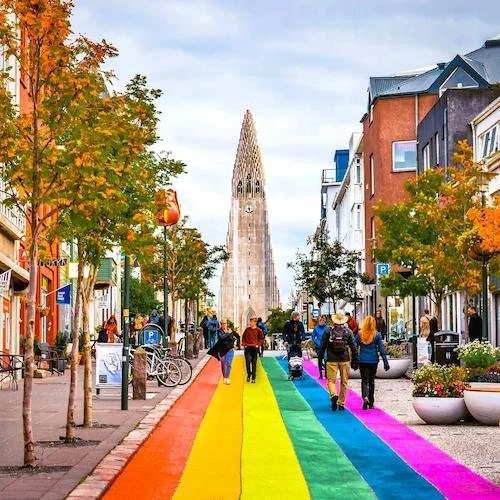 People walking on a rainbow-colored street in a festive outdoor area with colorful buildings and autumn trees.