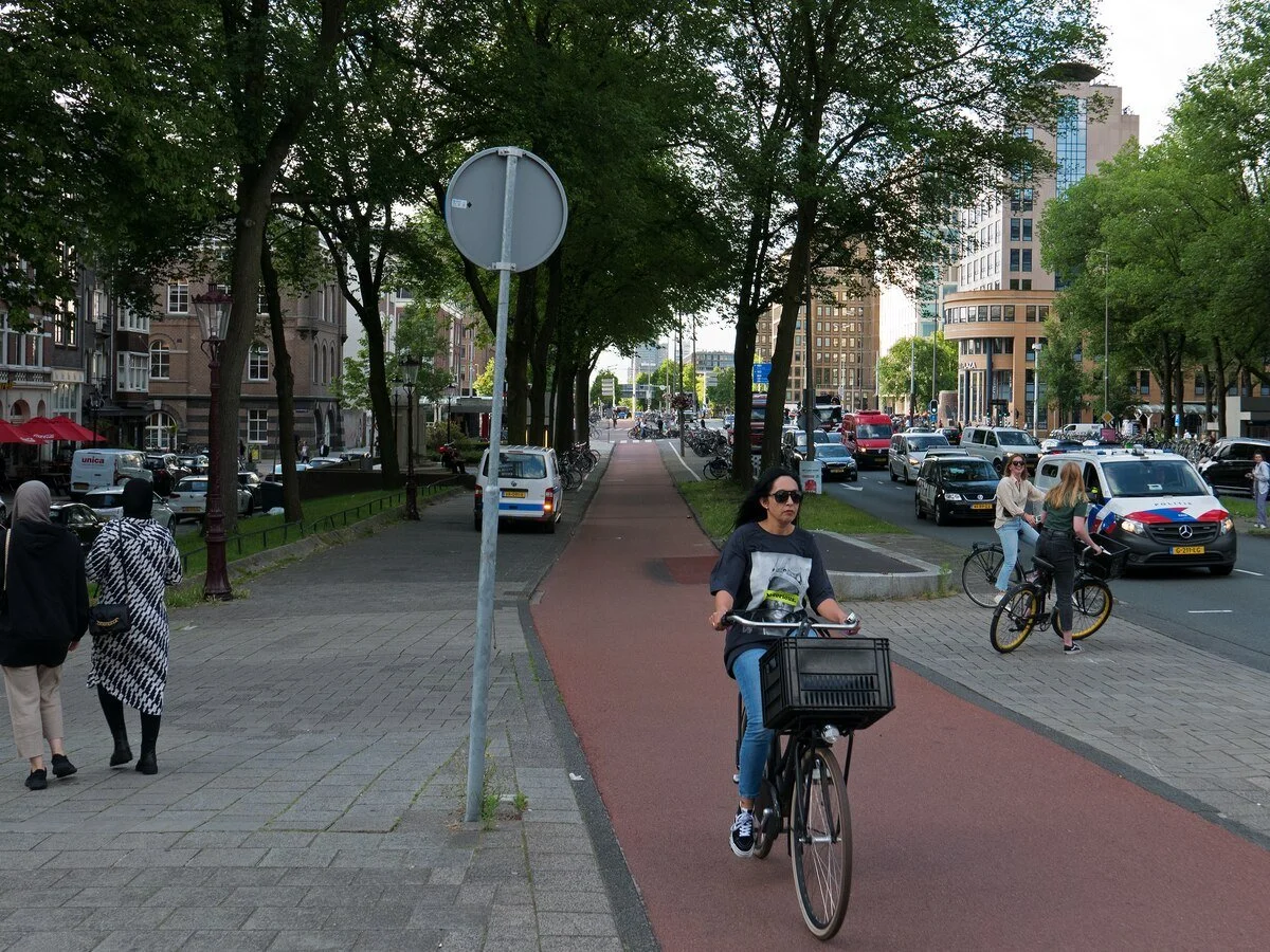 People walking and cycling on a city sidewalk and bike lane, with cars on the street and tall buildings in the background.