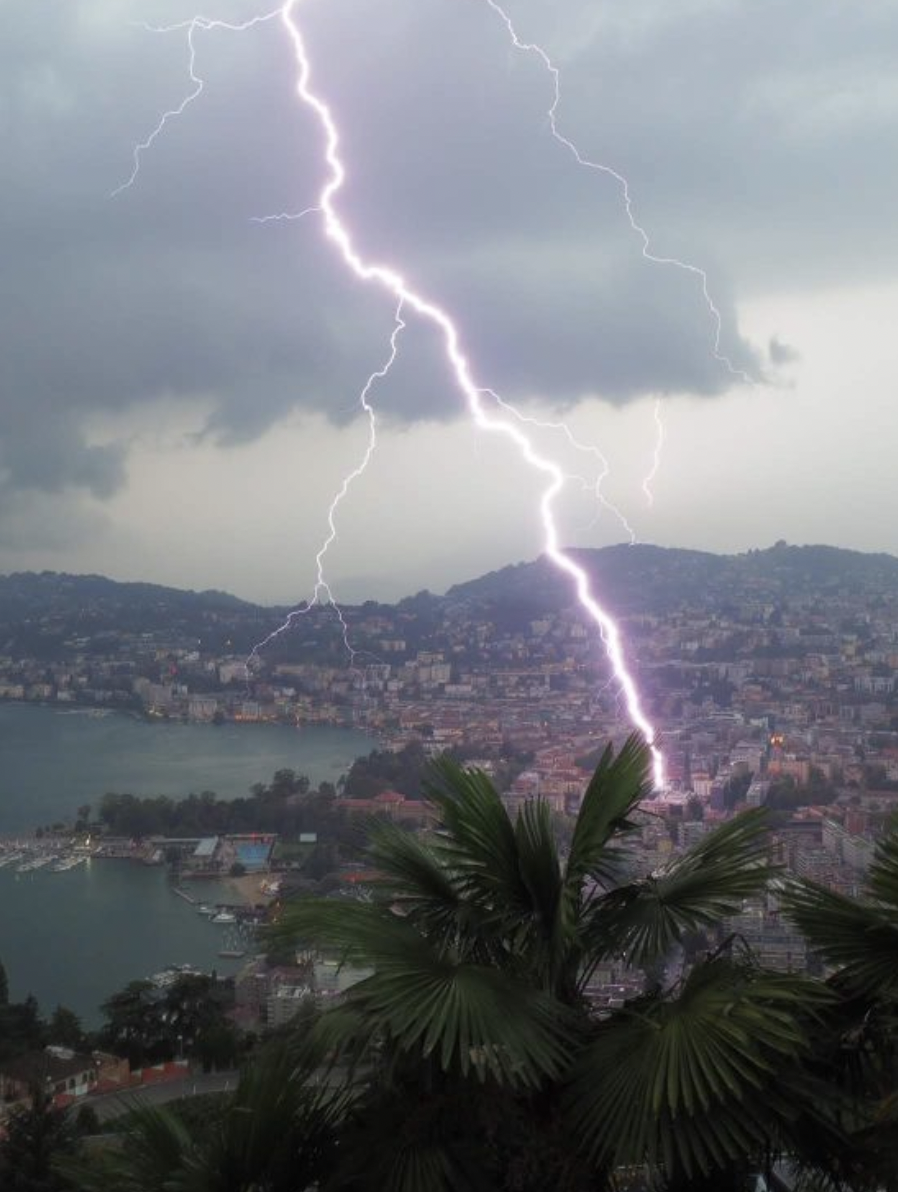 Lightning strikes over a city with hills, a body of water, and palm trees in the foreground, during a thunderstorm.