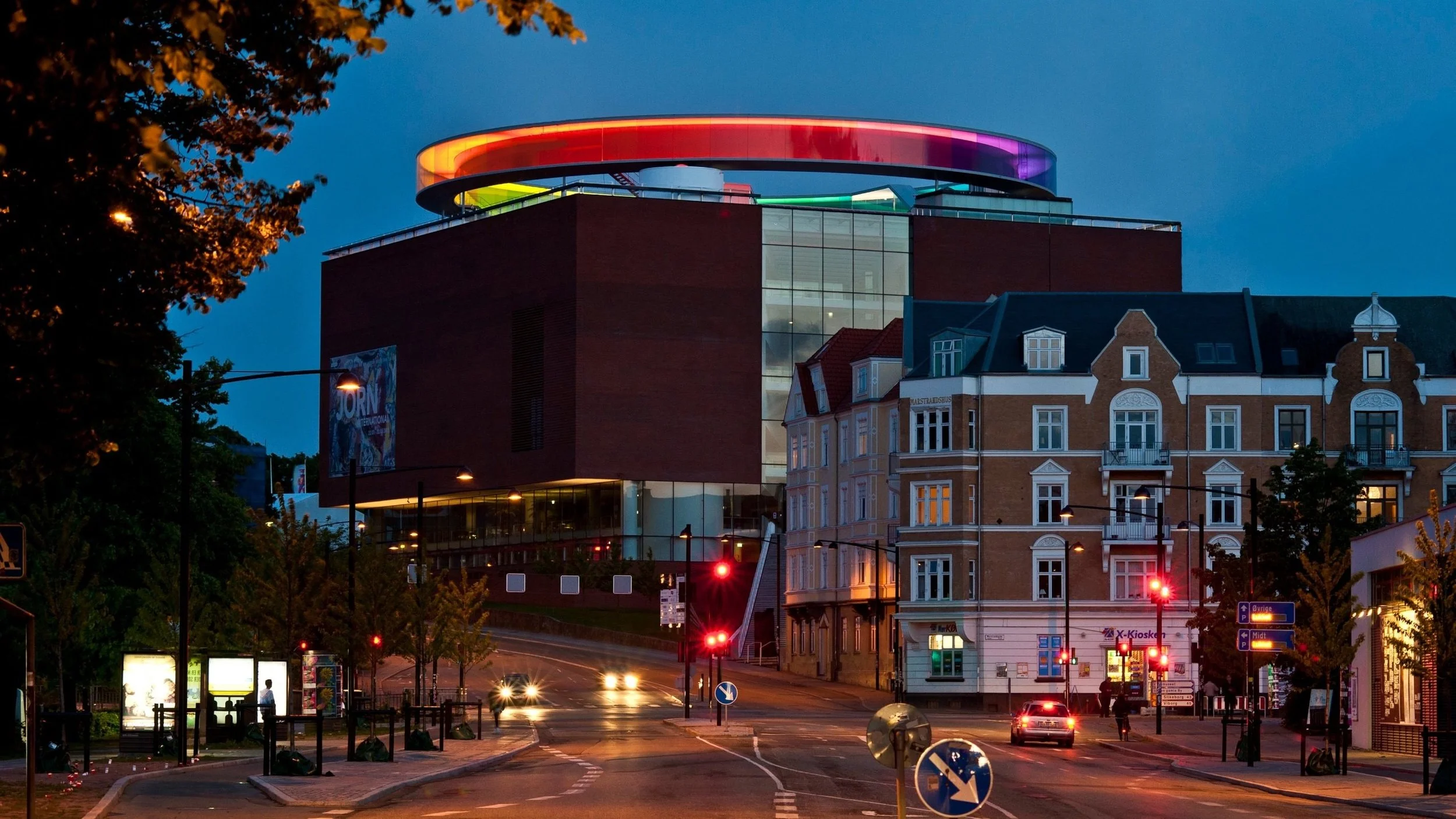 A city intersection at dusk with a modern building topped with a colorful illuminated ring and traditional European-style buildings nearby. Cars are driving through with their headlights on, and streetlights are lit along the streets.