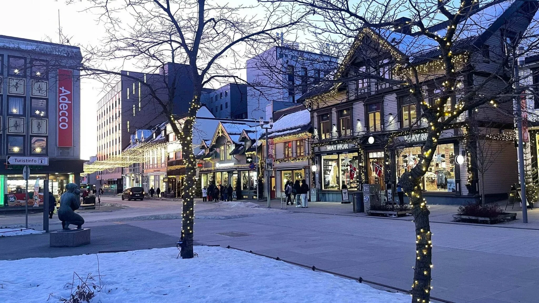 A winter street scene decorated with Christmas lights, with a snow-covered ground and several buildings with lit storefronts