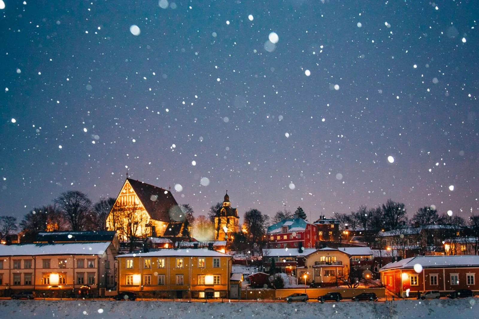 Snow falling over a small town during evening with illuminated church, houses, and trees on snow-covered ground.