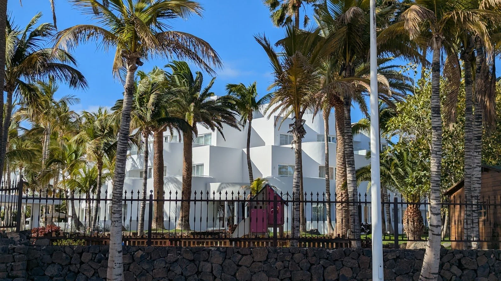Modern white apartment building surrounded by tall palm trees and a black iron fence, with a blue sky in the background.