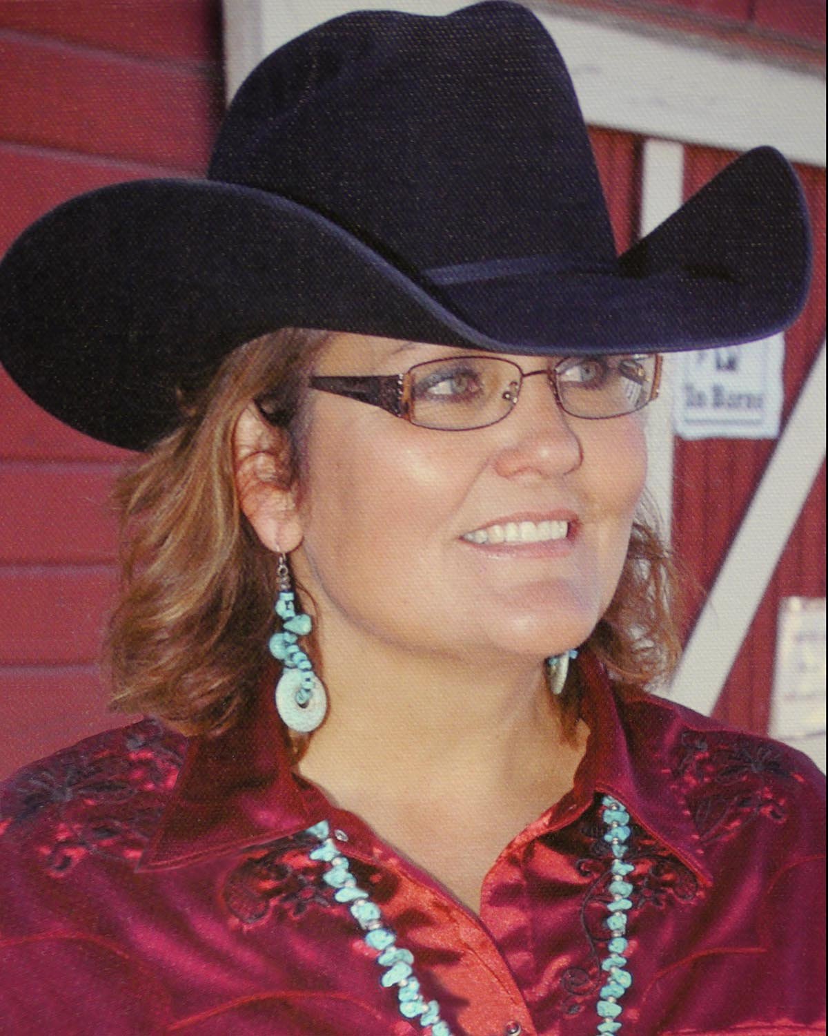 A woman wearing a black cowboy hat, glasses, turquoise jewelry, and a red satin Western shirt, smiling with a background of red wooden walls.