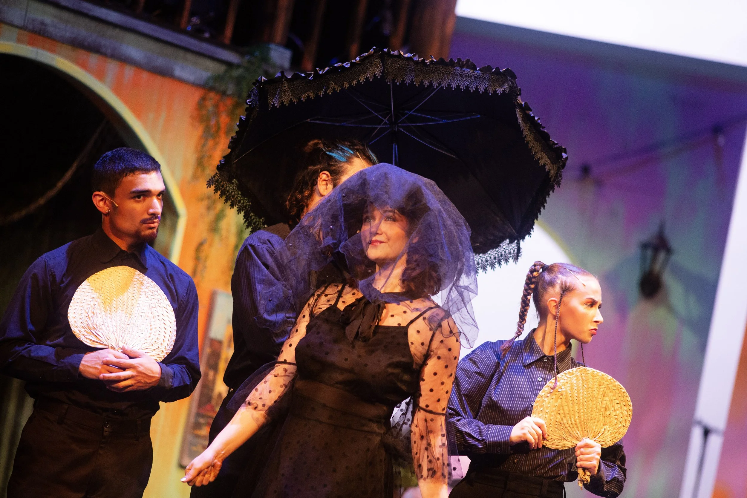 The photo shows four people stationed outdoors, with two women holding woven fans and another obscured woman holding an umbrella, engaged in a theatrical scene. The background is a building with an arched entrance, and the scene is lit with stage lig