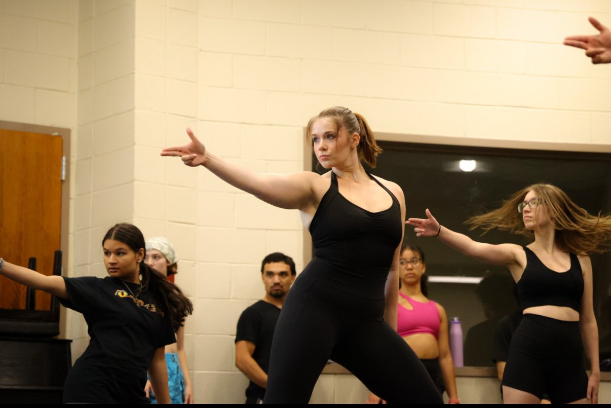 A group of young women participating in a dance or exercise class, with a woman in the center showing a pose with one arm extended, wearing athletic black clothing, in a classroom or studio setting.