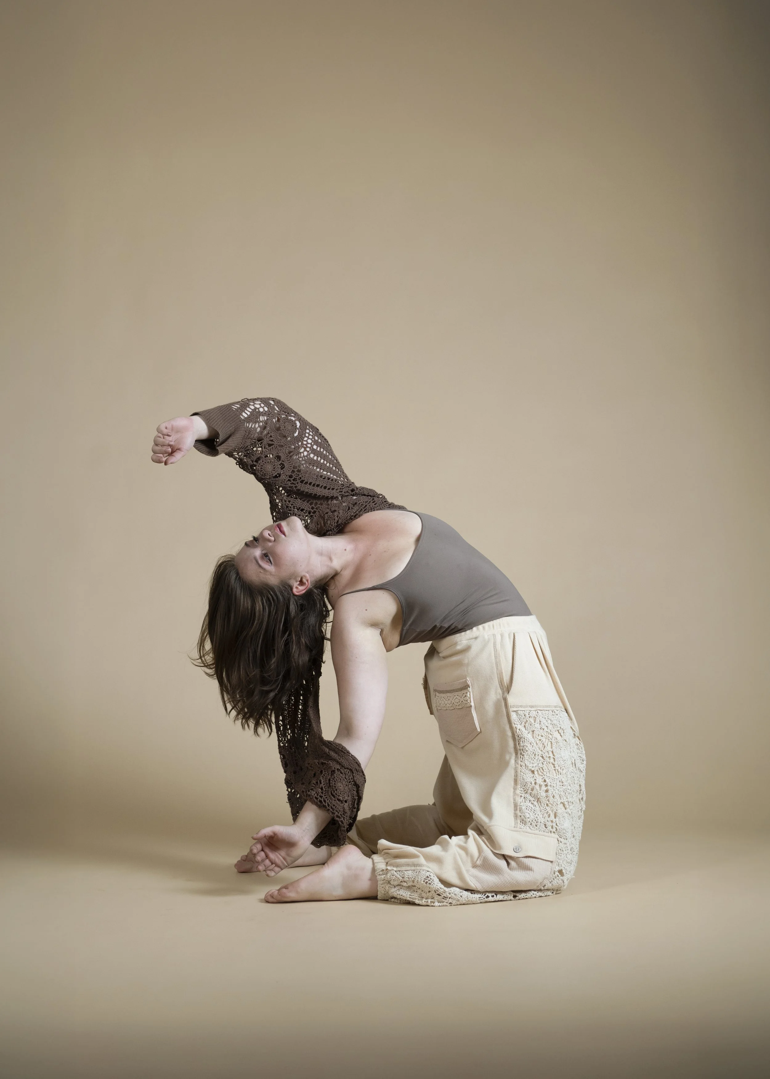 A woman with brown hair in a yoga pose on a beige background, with her body bent back and one arm extended, wearing a gray tank top and cream-colored pants with lace details.