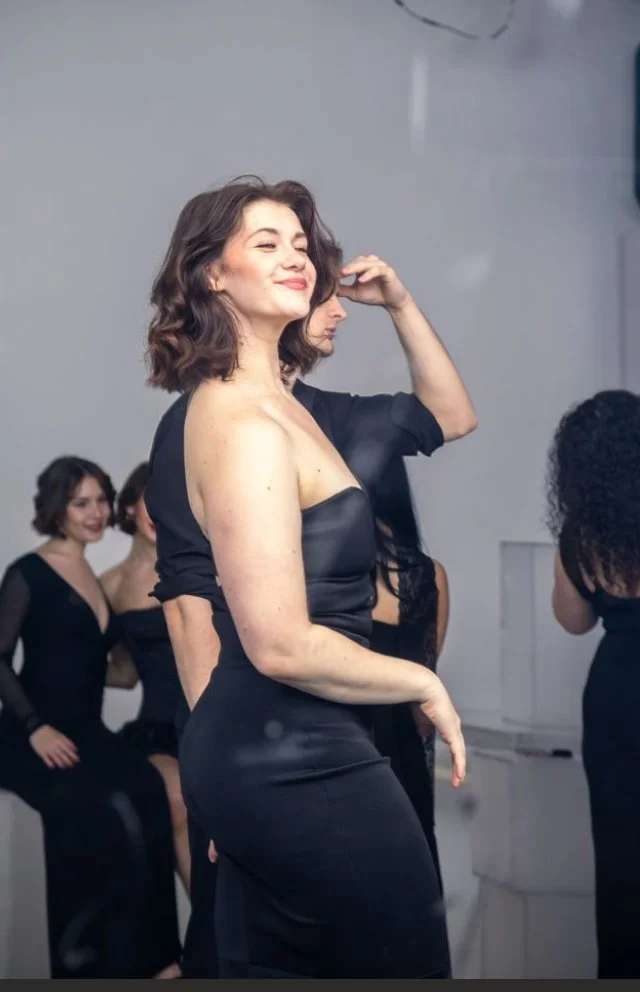 A woman with short, wavy brown hair posing confidently in a black dress, surrounded by others also dressed in black, against a plain indoor background.