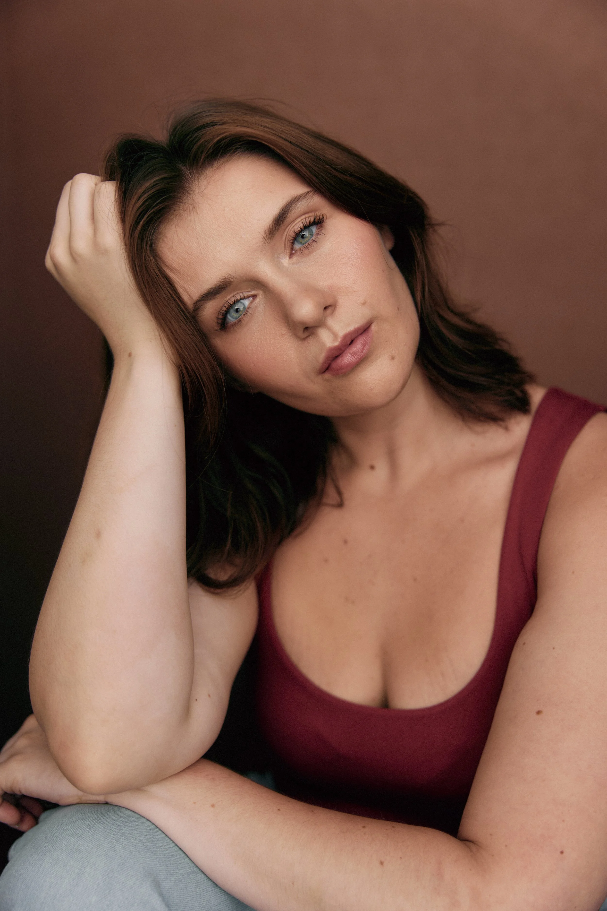 A woman with brown hair and blue eyes posing with her hand resting on her head against a brown background.