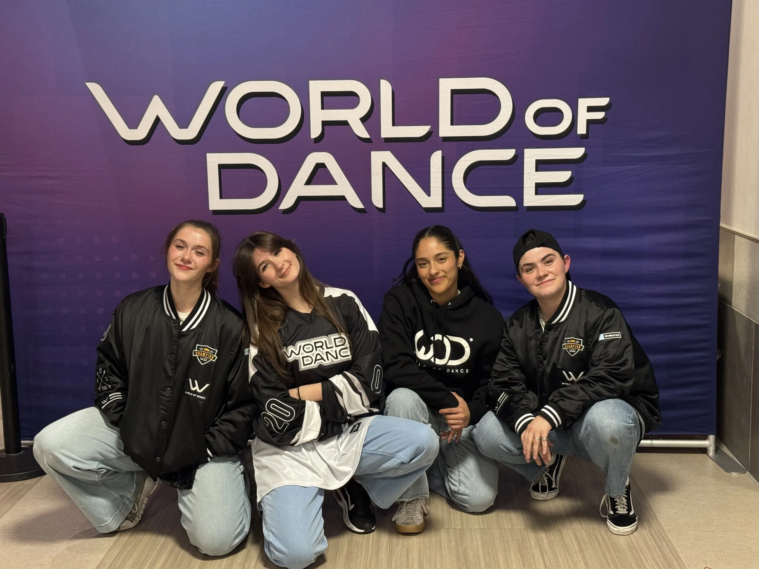 Four young dancers kneeling and smiling in front of a purple backdrop that reads 'WORLDOF DANCE', wearing casual black and sports jackets.