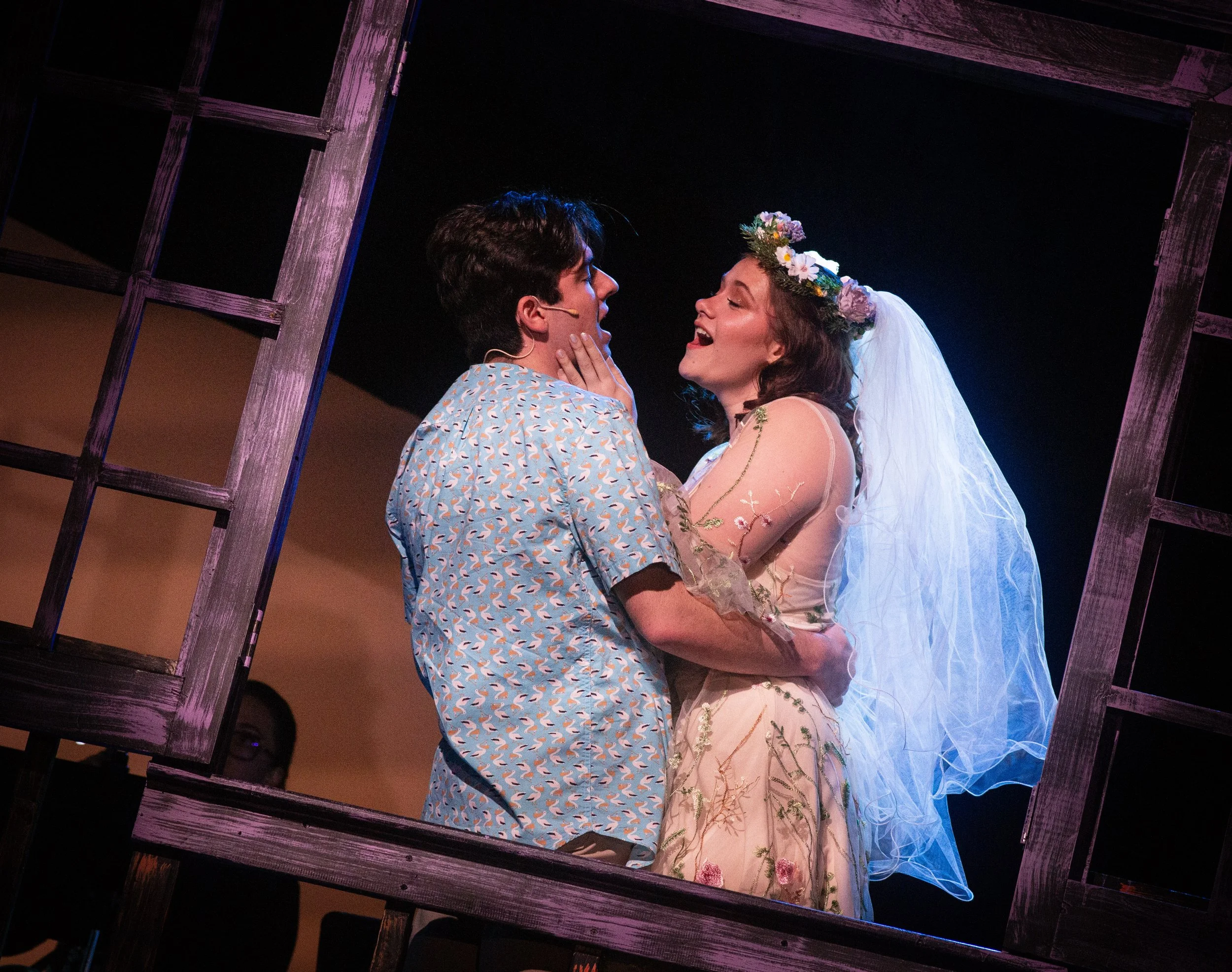A theatrical scene of a man and woman in wedding costumes, with the woman wearing a floral crown and veil, performing on stage framed by a wooden window prop.
