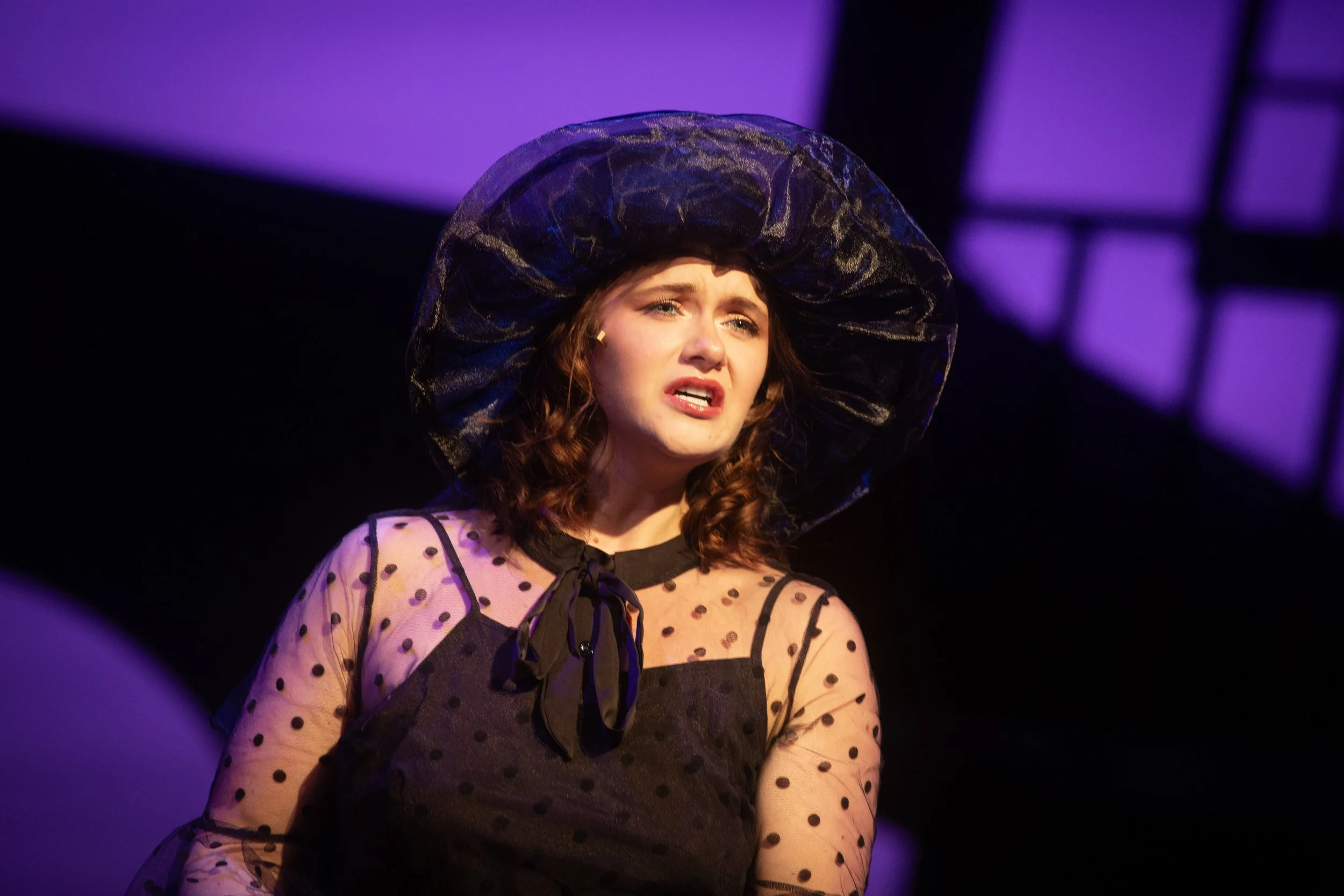 A woman with curly hair wearing a large black hat and a black polka dot dress performing on stage.