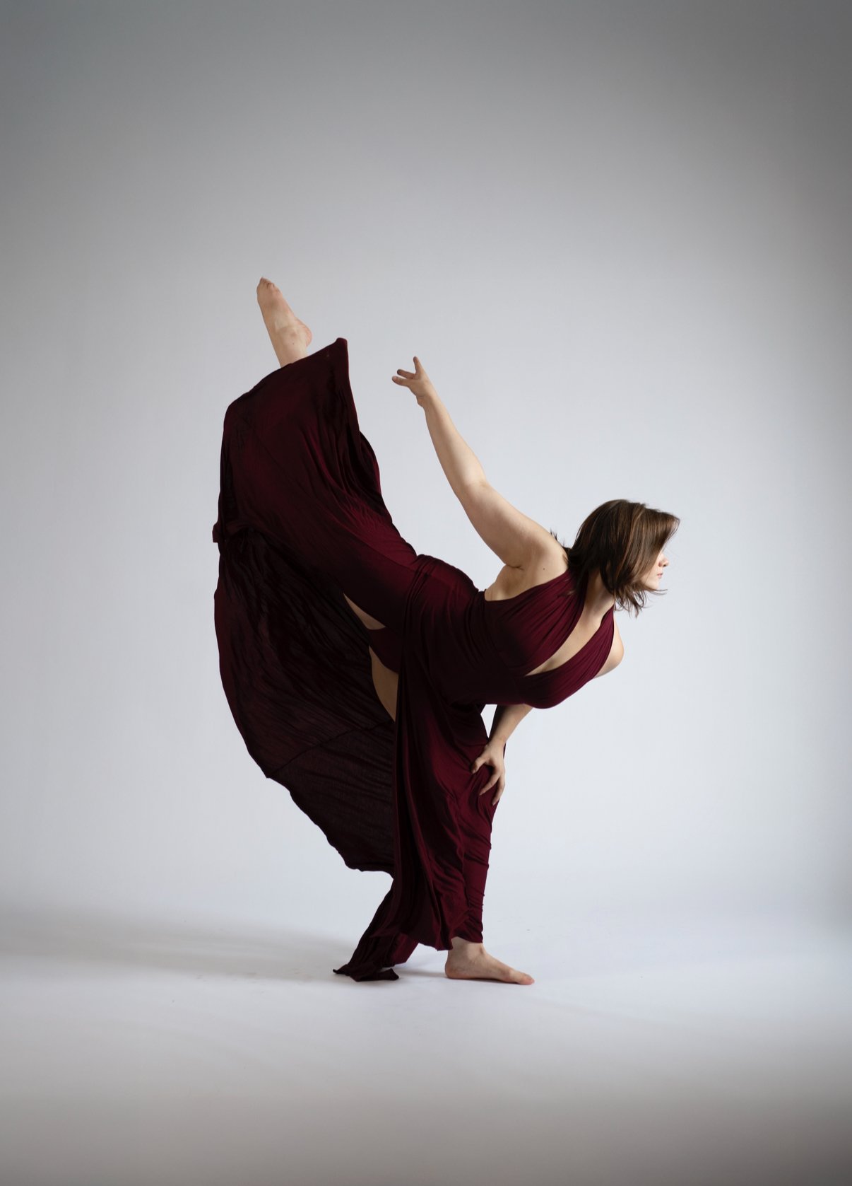 A woman dressed in a flowing burgundy dress performing a dance pose against a plain grey background.