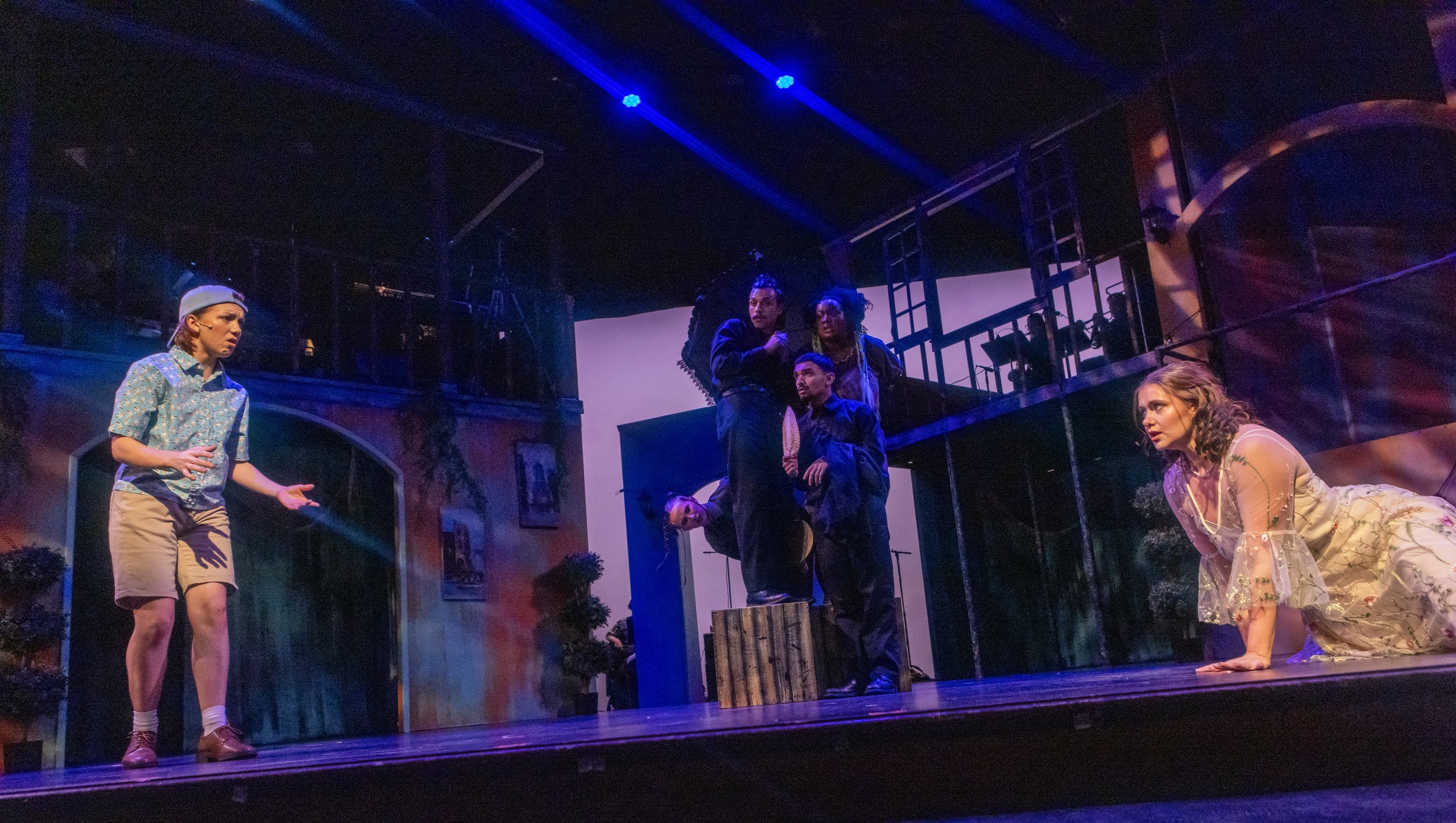 The scene is from a theatrical play with five actors on stage. The background features a multi-level set resembling a house interior with staircase and balcony. One actor on the left is standing with a puzzled expression, wearing shorts, a short-slee