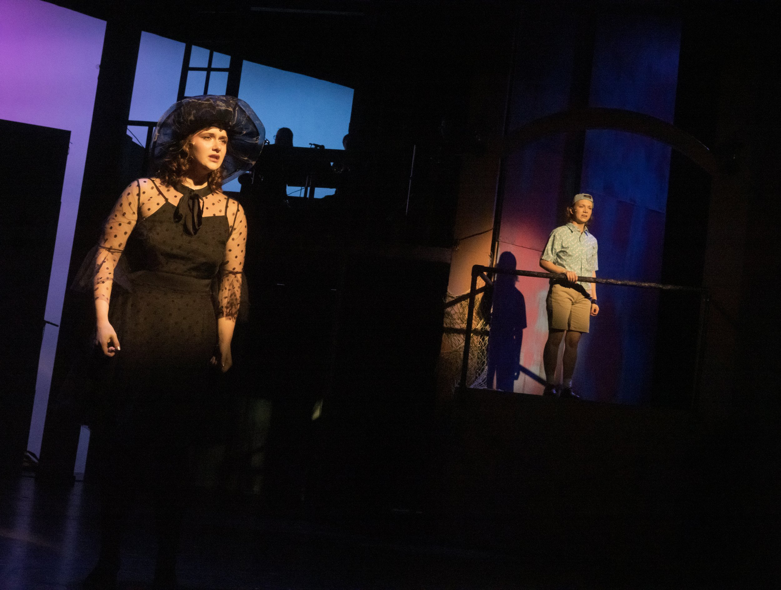 Two women on stage performing in a theater. One is dressed in a black polka dot dress with sheer sleeves and a large hat, looking emotional. The other is in a casual shirt and shorts, standing on a balcony, with a more neutral expression.