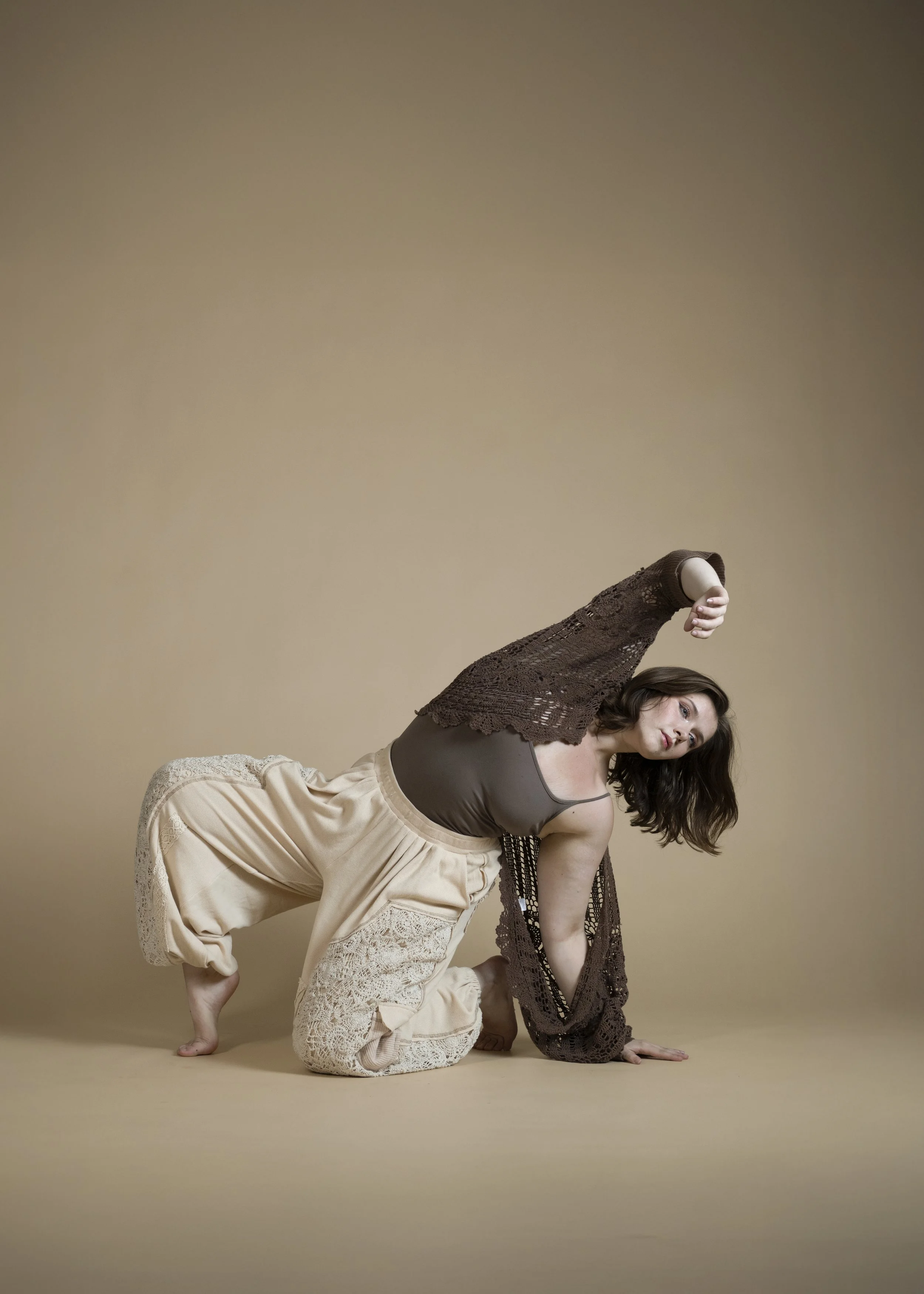 A woman with dark hair posing on a beige background, wearing loose beige pants, a brown tank top, and a brown lace cardigan, with one arm extended and one hand on the ground.
