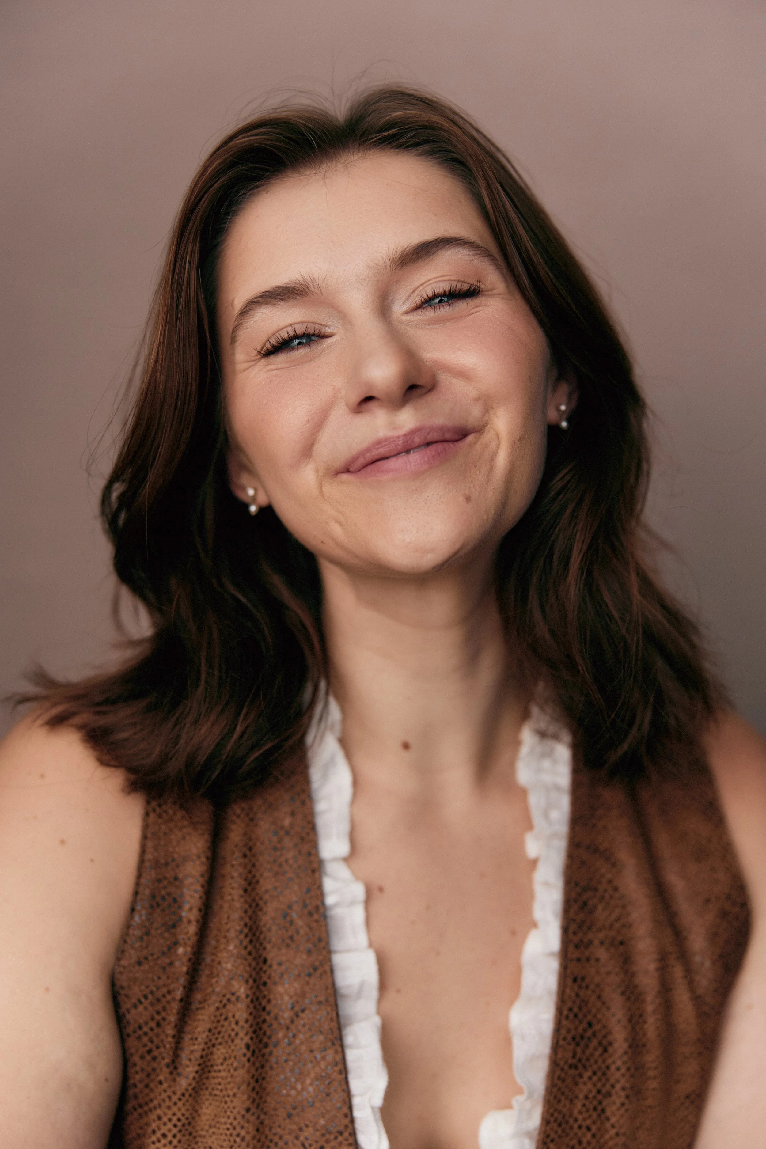 Close-up of a young woman with shoulder-length brown hair, smiling with eyes partially closed, wearing a sleeveless brown top with white ruffled detail and small earrings.