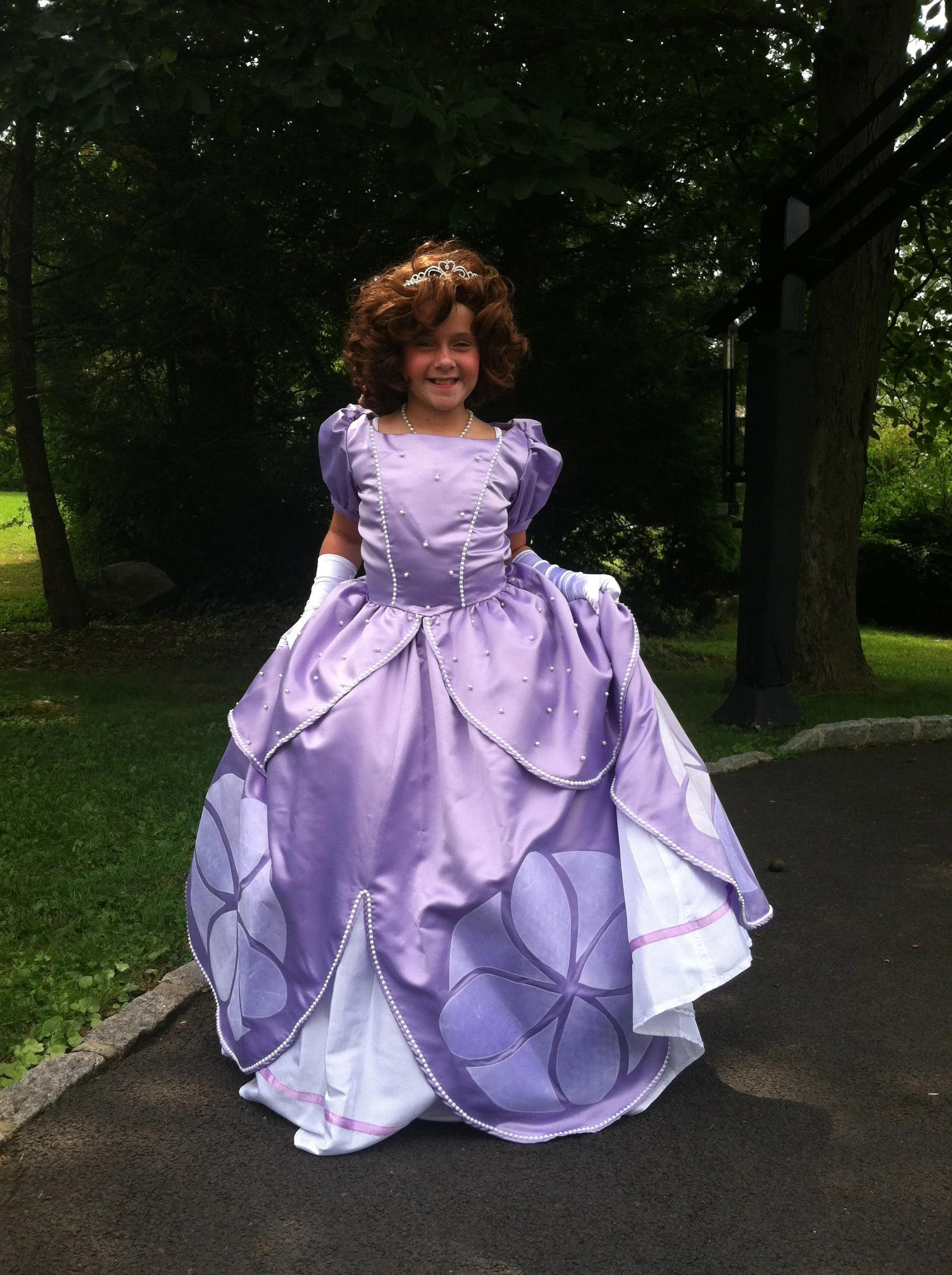 Young girl dressed in a lavender princess gown with pearl accents, wearing a tiara, standing outdoors on a paved path surrounded by trees and greenery.