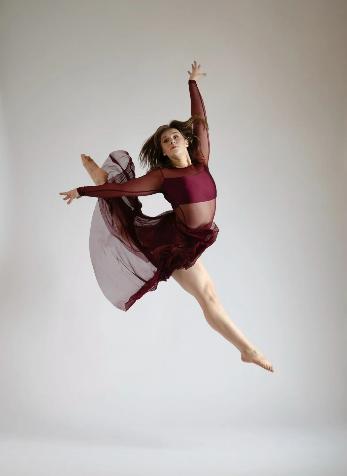A woman in a maroon dance costume performing a jump with her arms raised and one leg extended outward.