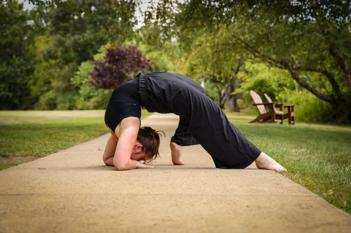 A woman dancing in a park, performing a pose on a paved pathway surrounded by trees and greenery.