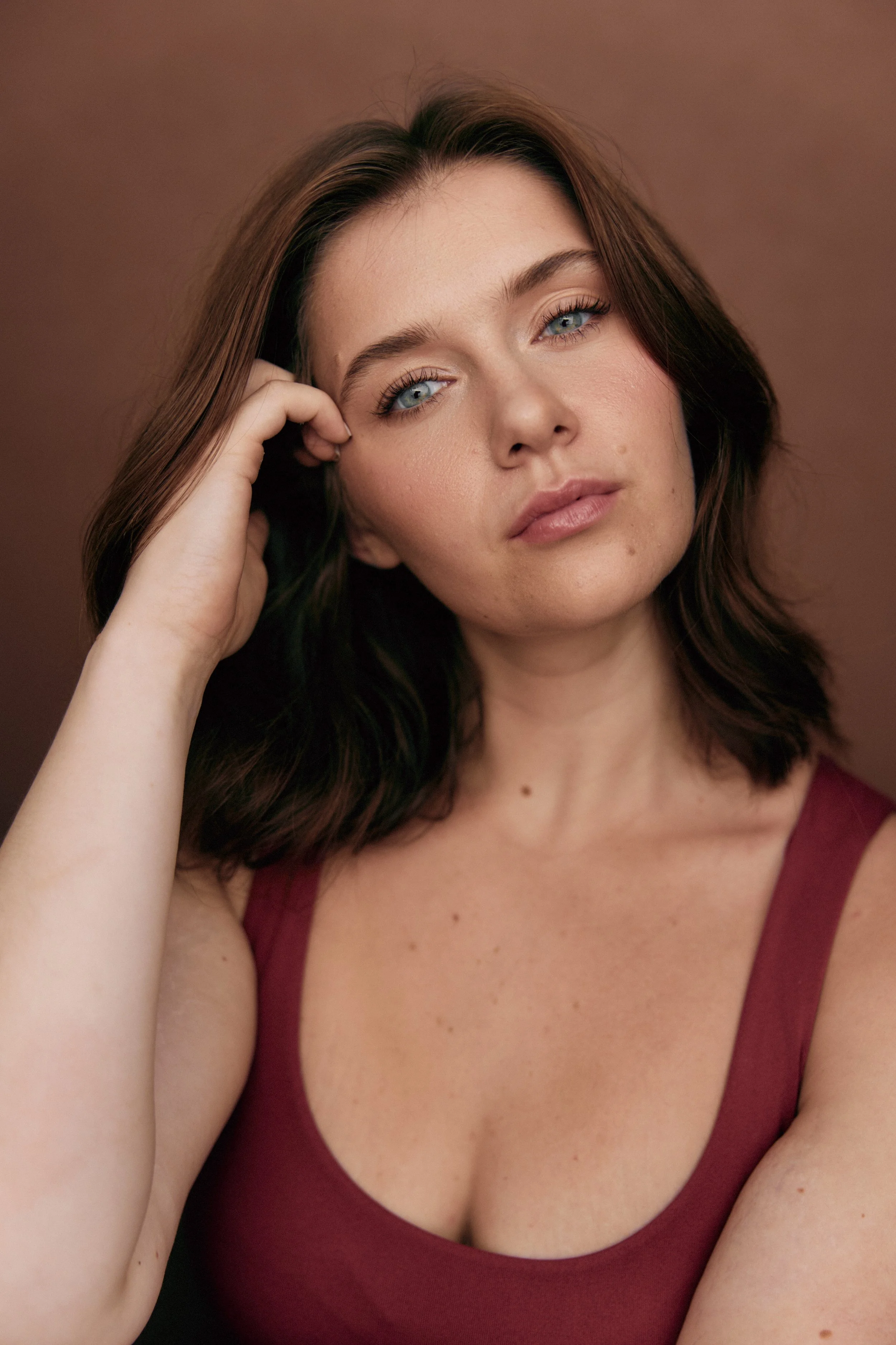 Close-up of a woman with brown hair and blue eyes, wearing a maroon sleeveless top, resting her head on her hand, against a brown background.