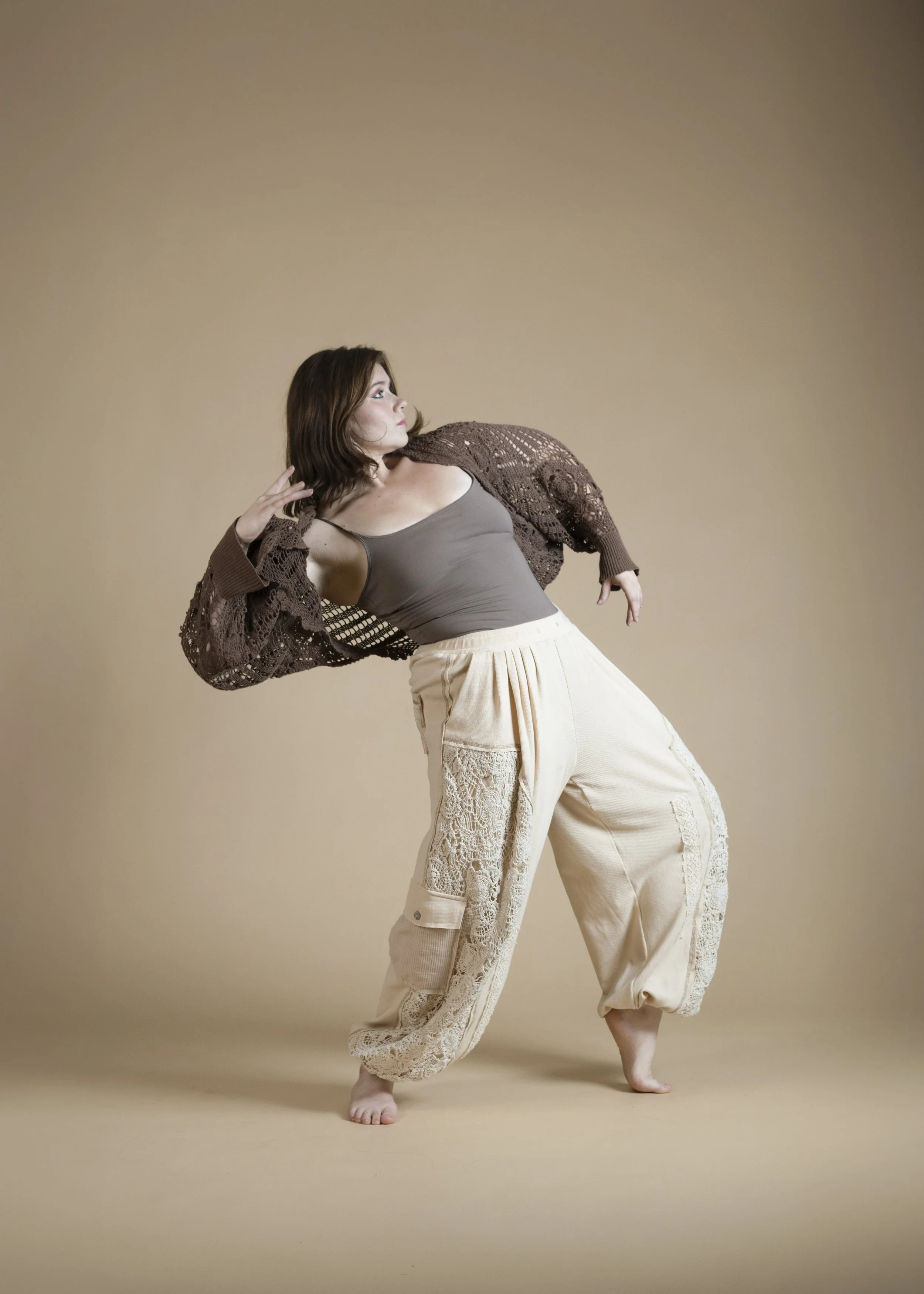 A woman with shoulder-length brown hair posing against a beige background, wearing a gray tank top, beige pants with lace details, and a brown crocheted cardigan.