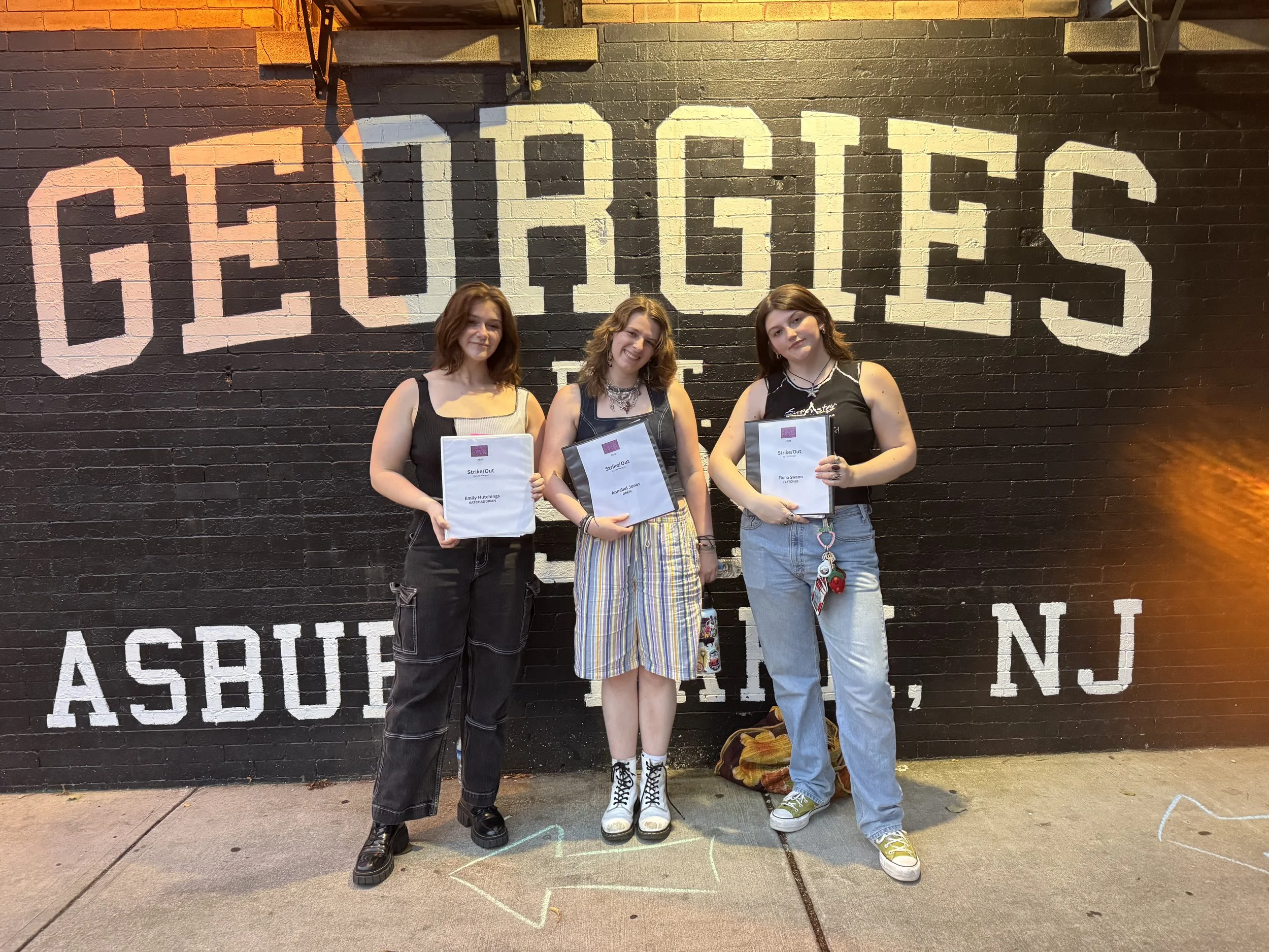 Three young women standing in front of a black brick wall with 'GEORGETOWN' painted on it in large white letters, each holding a play script. The wall also has 'ASBURY' and 'NJ' painted at the bottom. They are smiling and dressed casually.