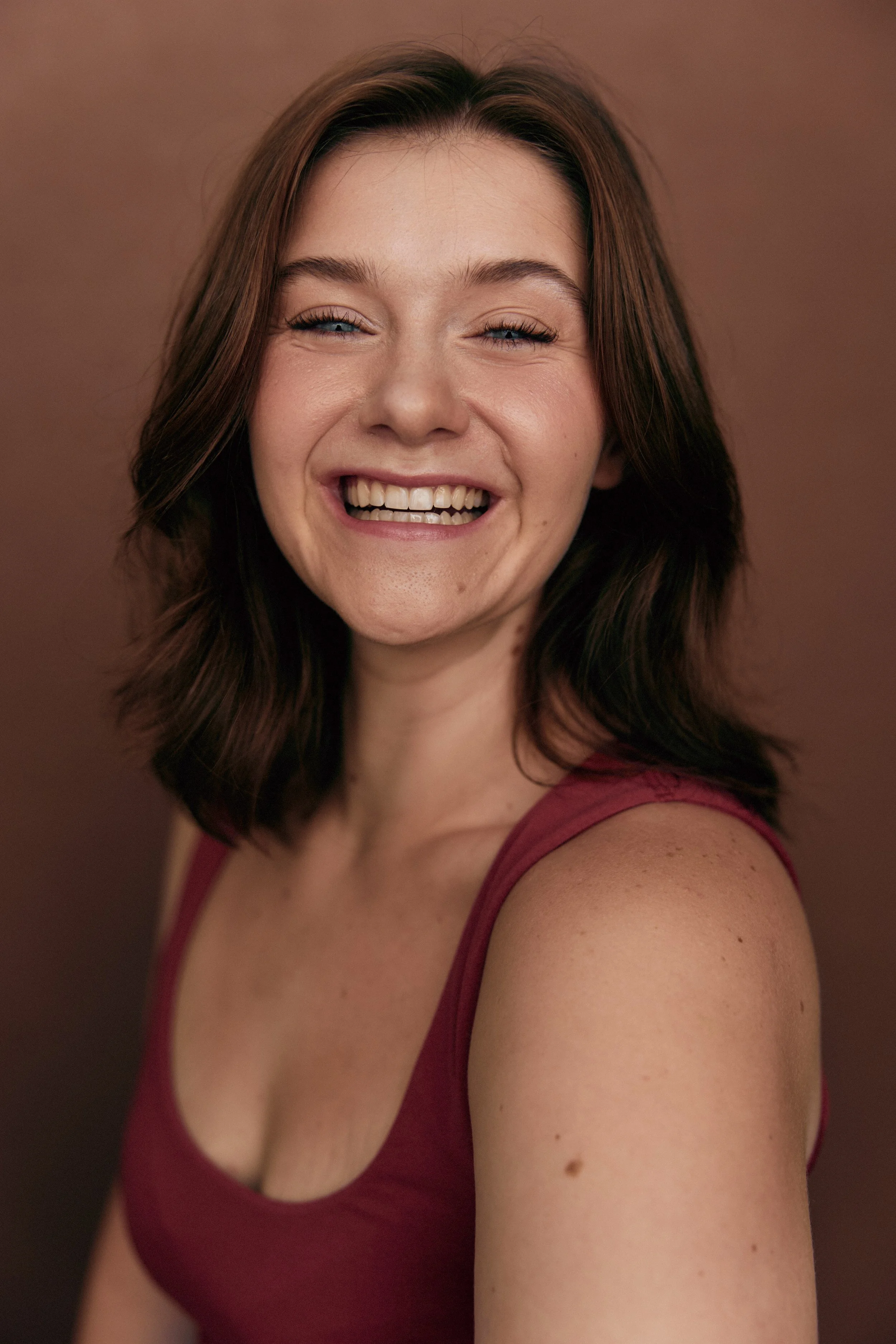 A woman with shoulder-length brown hair smiling showing her teeth, wearing a sleeveless maroon top, against a plain brown background.