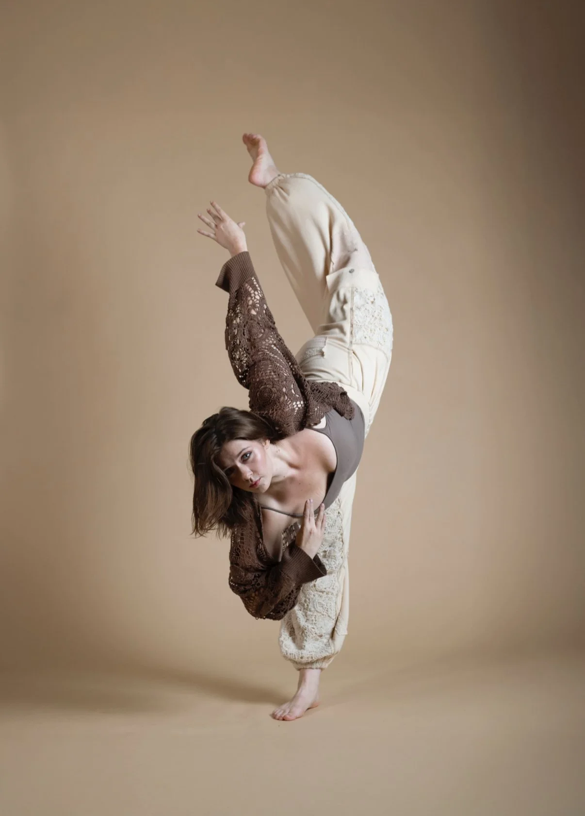 A woman performing a one-armed handstand or contortion move against a beige background, wearing loose cream-colored pants, a gray top, and a brown lace cardigan.