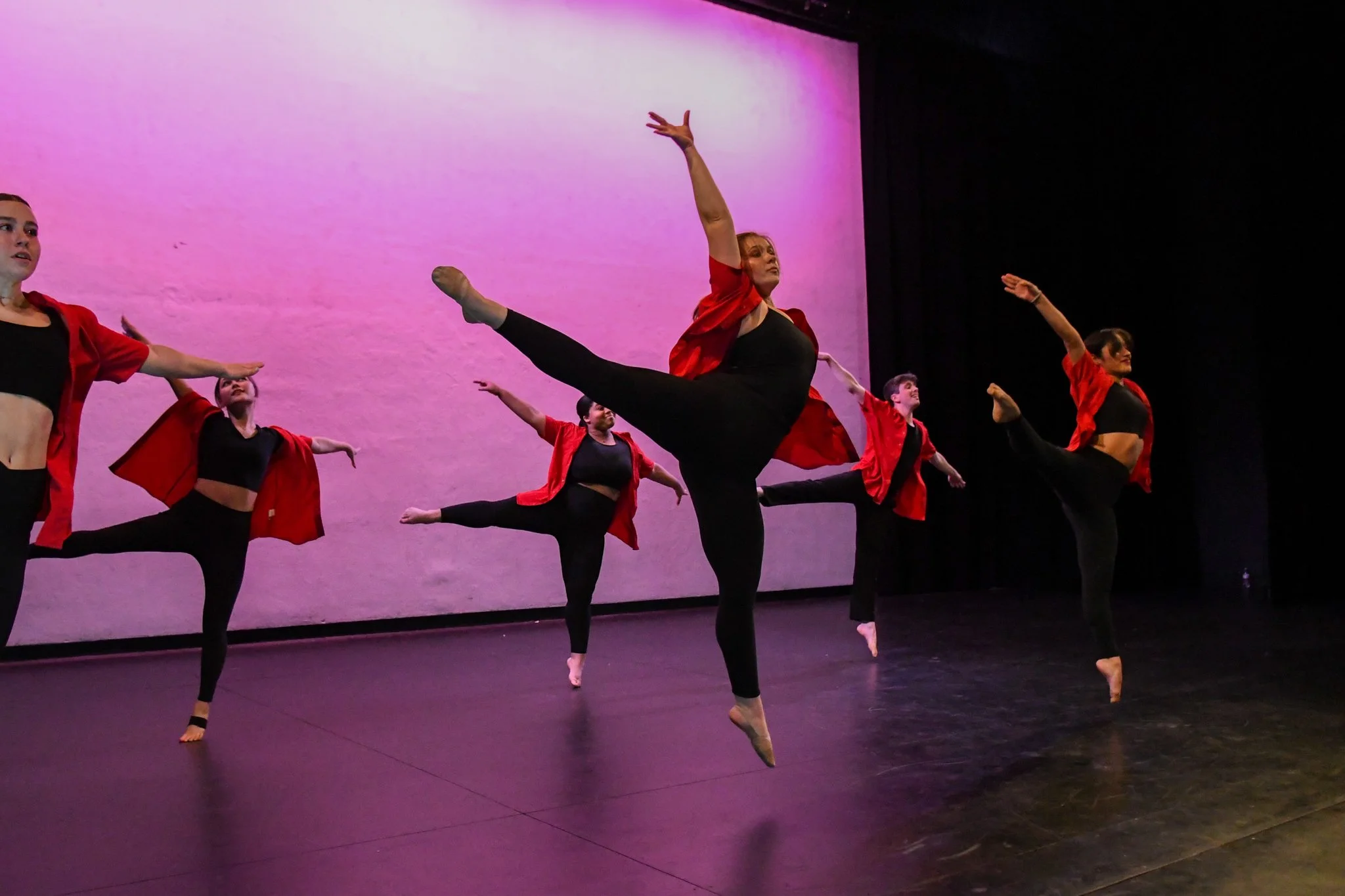 Group of five dancers performing on stage, mid-air, with purple background. They wear black outfits with red jackets, dancers are in various leap positions.
