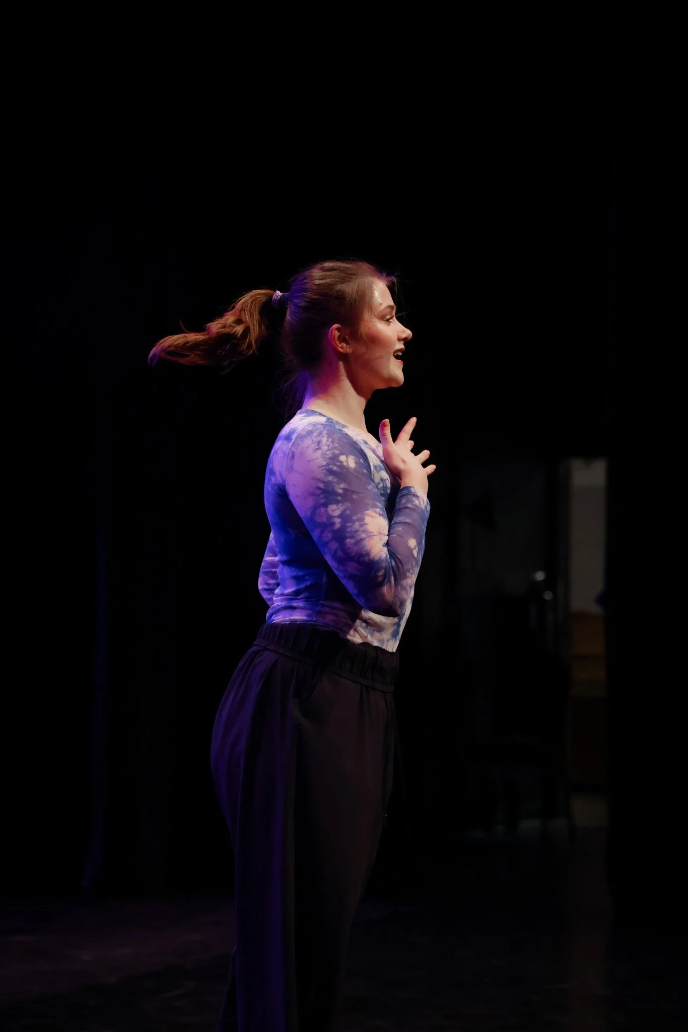 A woman with a ponytail and a tie-dye long sleeve shirt standing on stage, smiling, with her right hand near her face.