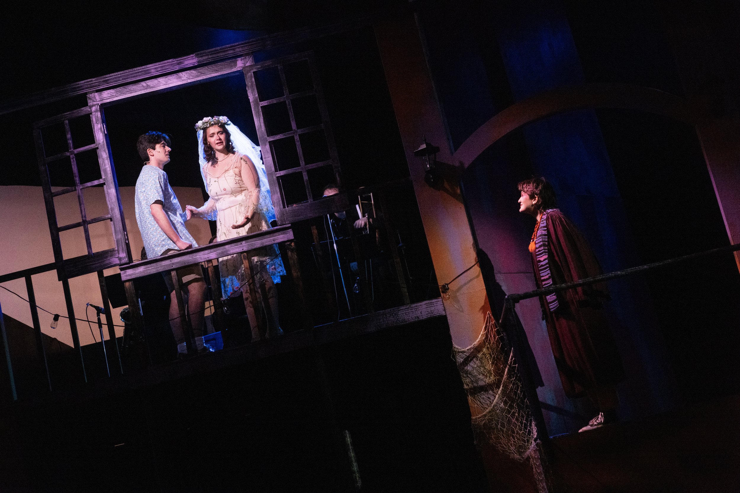 Scene from a theatrical play with three actors on stage, two of them behind a window frame, one in front of it, in a dimly lit setting with dark and colorful background.