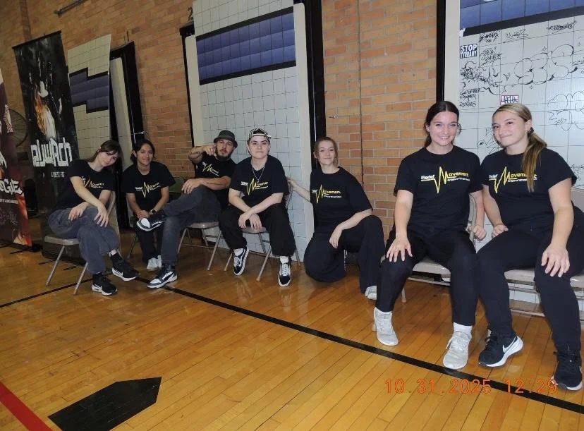 Seven people sitting on chairs in a gymnasium, wearing black t-shirts with a yellow heartbeat line and text, with a brick wall and whiteboard behind them.