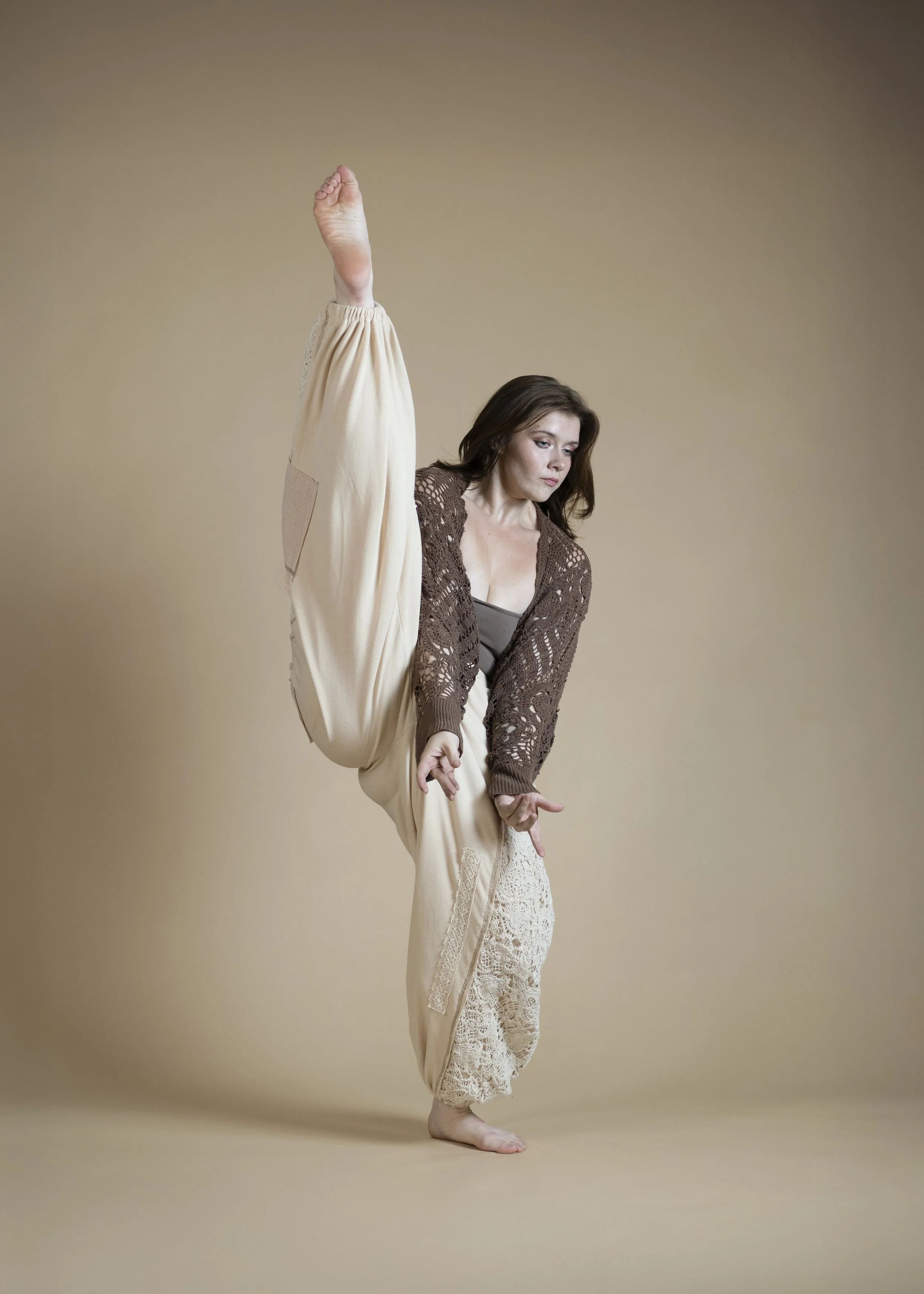 A woman standing on one leg with her other leg raised high, holding her foot with one hand while extending her other arm downward, dressed in loose, comfortable clothing, in a studio with a beige background.