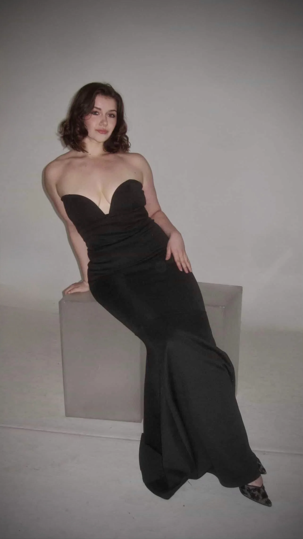 A woman in a strapless black gown sitting on a gray box against a plain wall.
