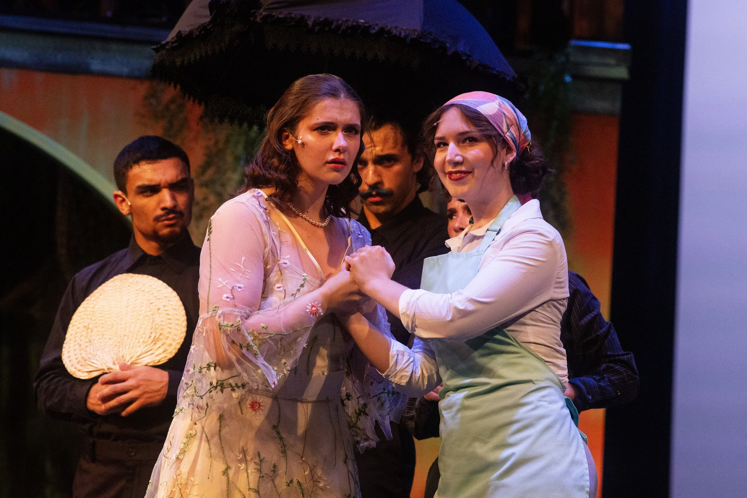 Four actors on stage performing in a theatrical scene. Two women are holding hands and facing each other, one in a floral dress and pearl necklace, the other in a white apron and headscarf, both with serious expressions. Two men are in the background