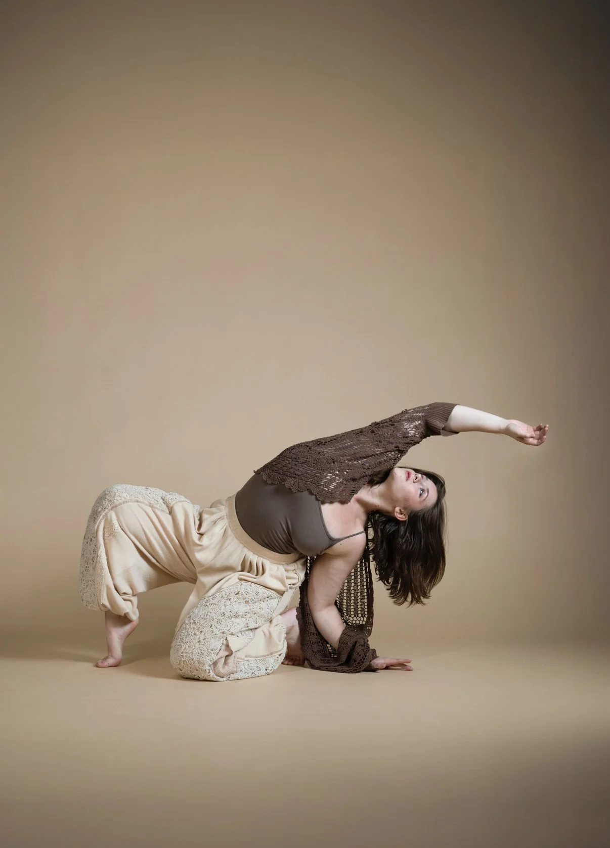 A woman performing a dance pose on a beige background, bent forward with one arm extended upward and the other arm supporting her on the ground, dressed in comfortable clothing.