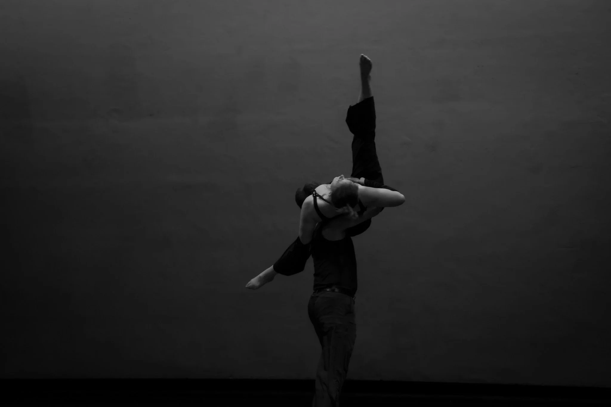 A ballet dancer is being lifted by a male performer on a dark stage, with the dancer's legs extended upwards and body bent at the waist.