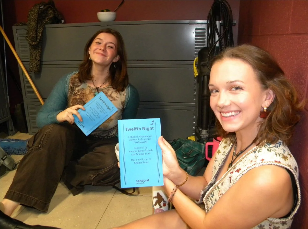 Two women sitting on the floor, smiling, holding scripts or programs for Twelfth Night, a musical adaptation of William Shakespeare's play. One woman has shoulder-length brown hair and is wearing a teal cardigan, the other has curly hair and is weari